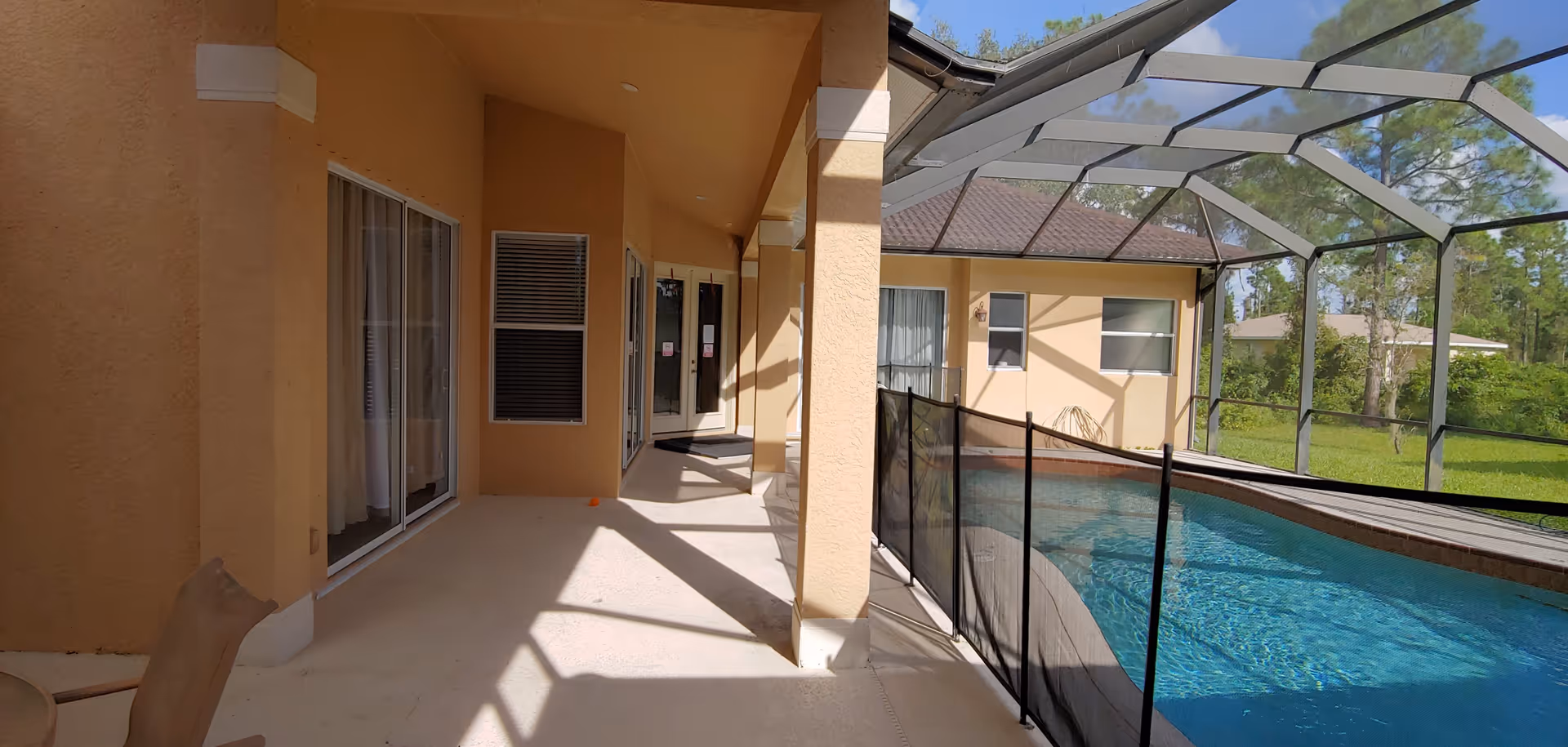 A covered outdoor patio area adjacent to a screened-in swimming pool with a safety fence. The patio has beige walls and a concrete floor, with sliding glass doors and windows leading into the building. Trees and greenery are visible outside the screened enclosure.