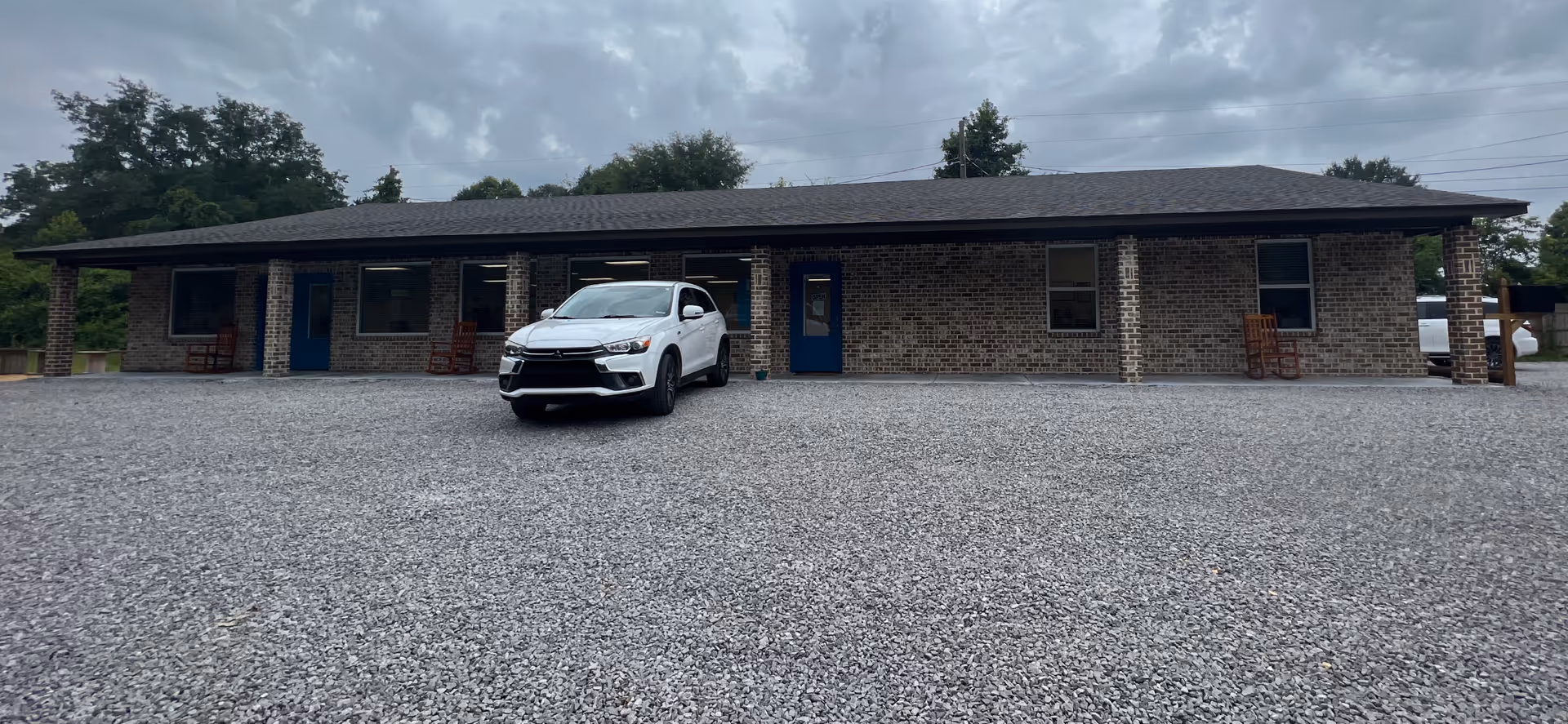Single-story brick building with blue doors, rocking chairs, and a white SUV in front.