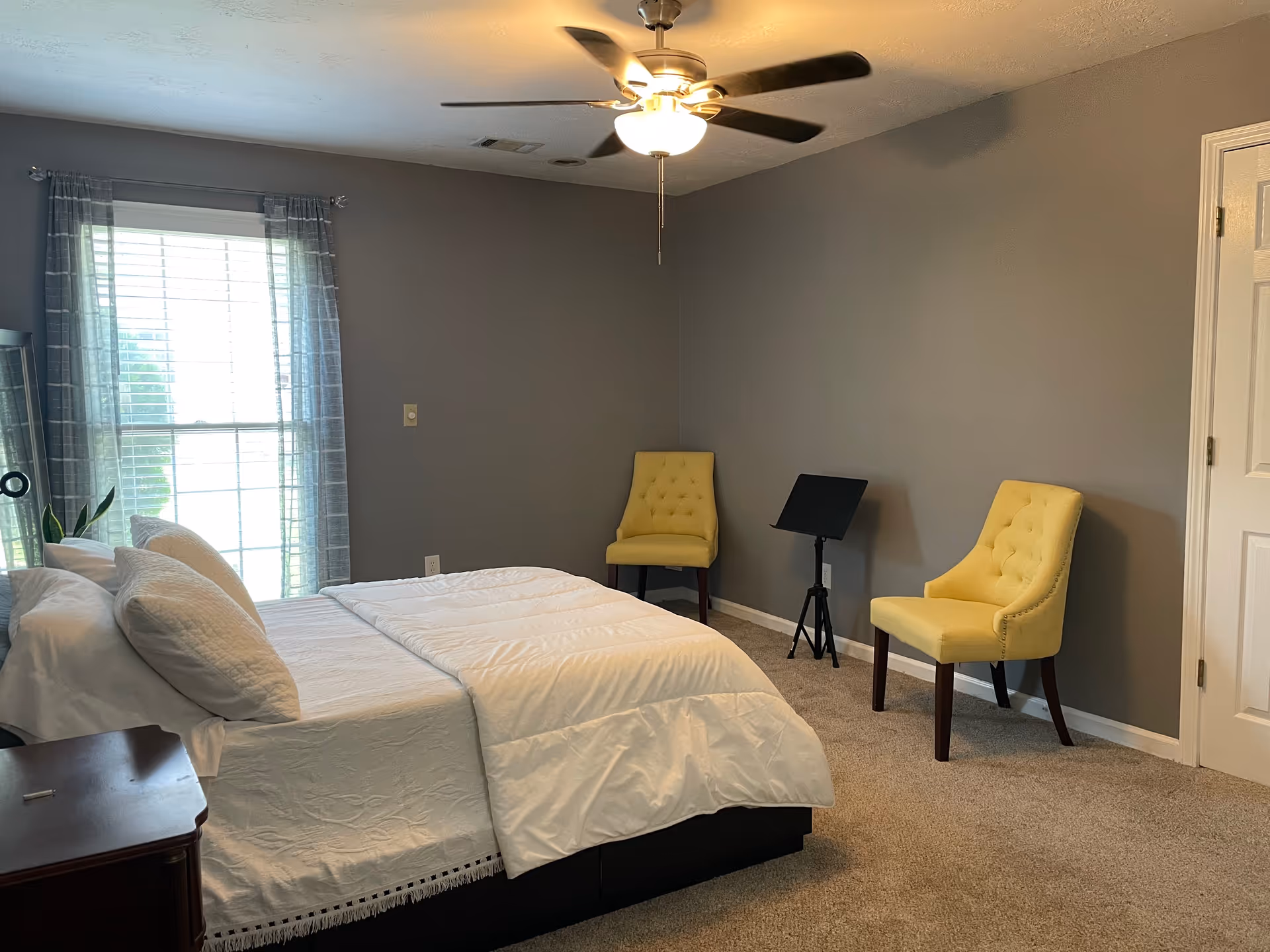 Furnished bedroom with a bed, two yellow chairs, ceiling fan, and window.