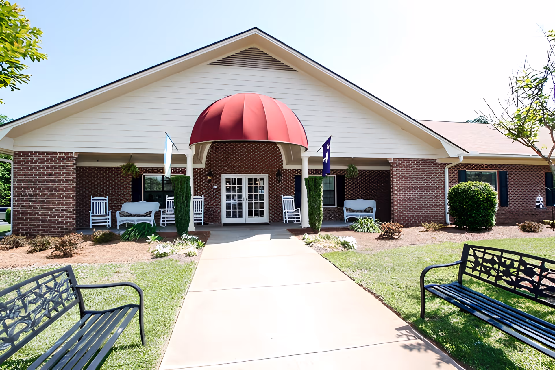Front entrance of a brick building with a red dome-shaped awning over double glass doors. The entrance is flanked by white rocking chairs and benches on a covered porch. Two black metal benches are positioned on either side of a concrete walkway leading to the door. The surrounding area has green grass, small shrubs, and trees under a clear blue sky.
