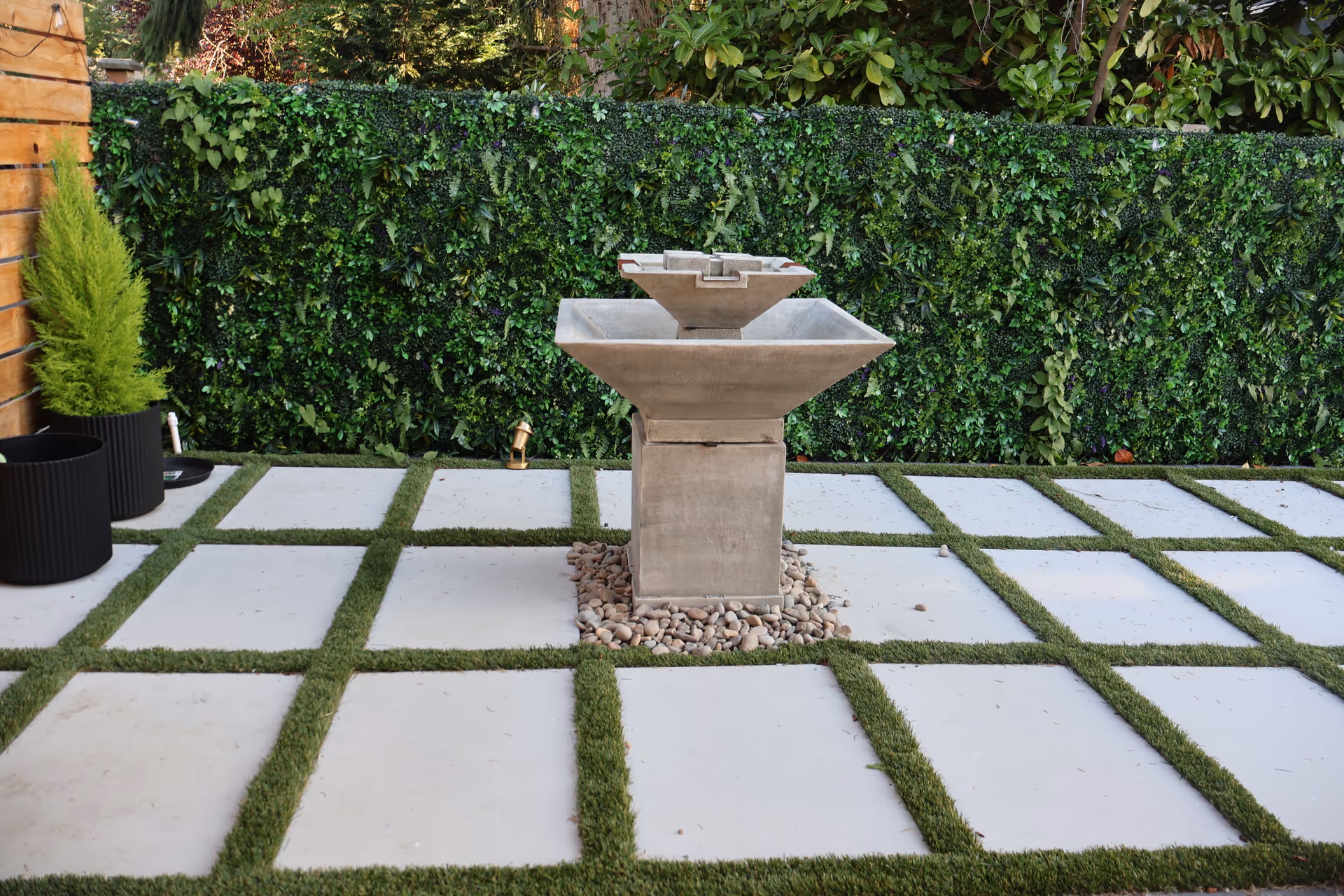 A modern stone fountain sits on a paved courtyard with greenery and a hedge.