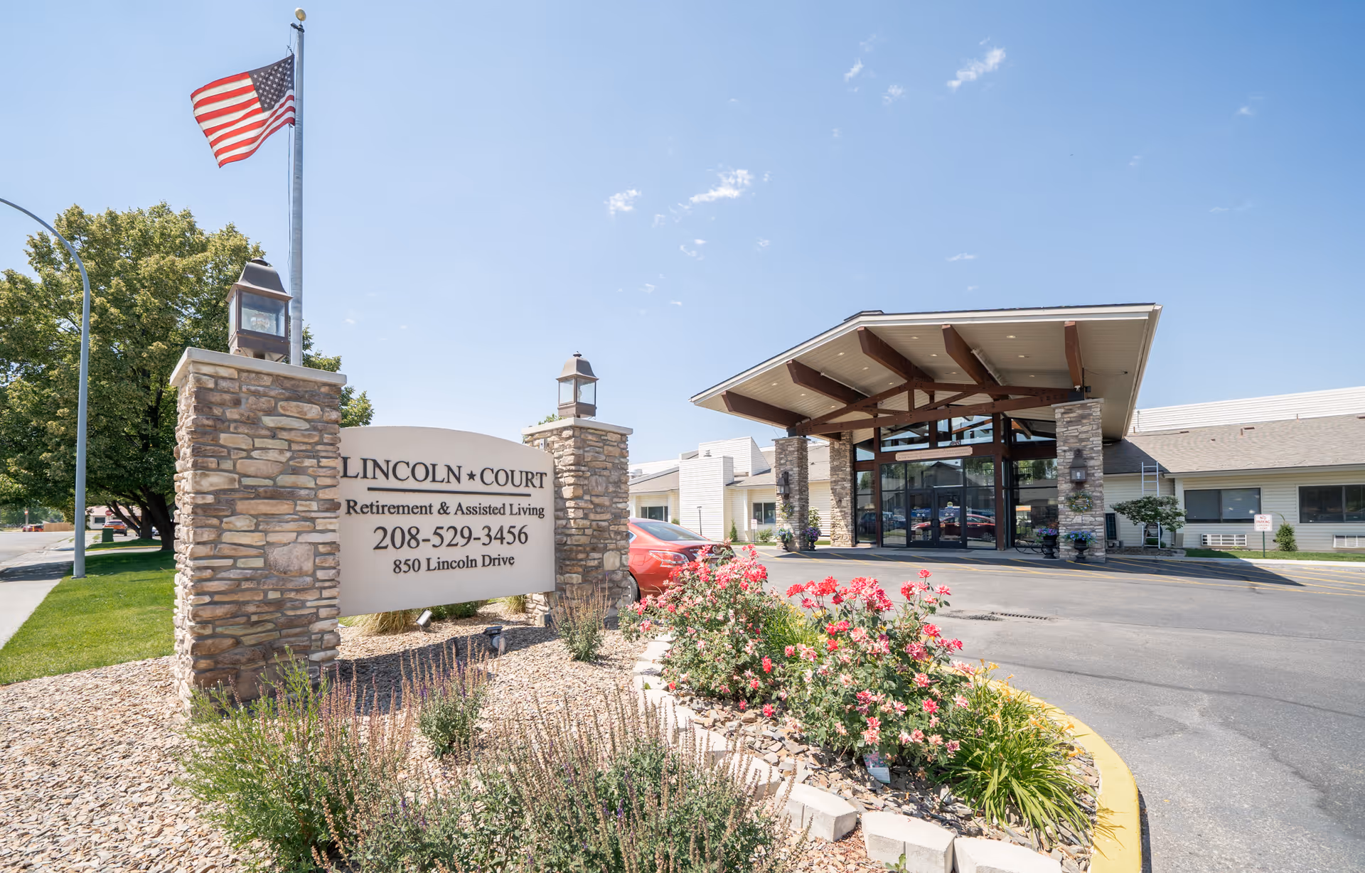 Exterior view of Lincoln Court Assisted & Senior Living facility showing the entrance with a covered drop-off area, stone pillars, landscaped flower beds, an American flag on a flagpole, and a sign with the facility name, phone number, and address.