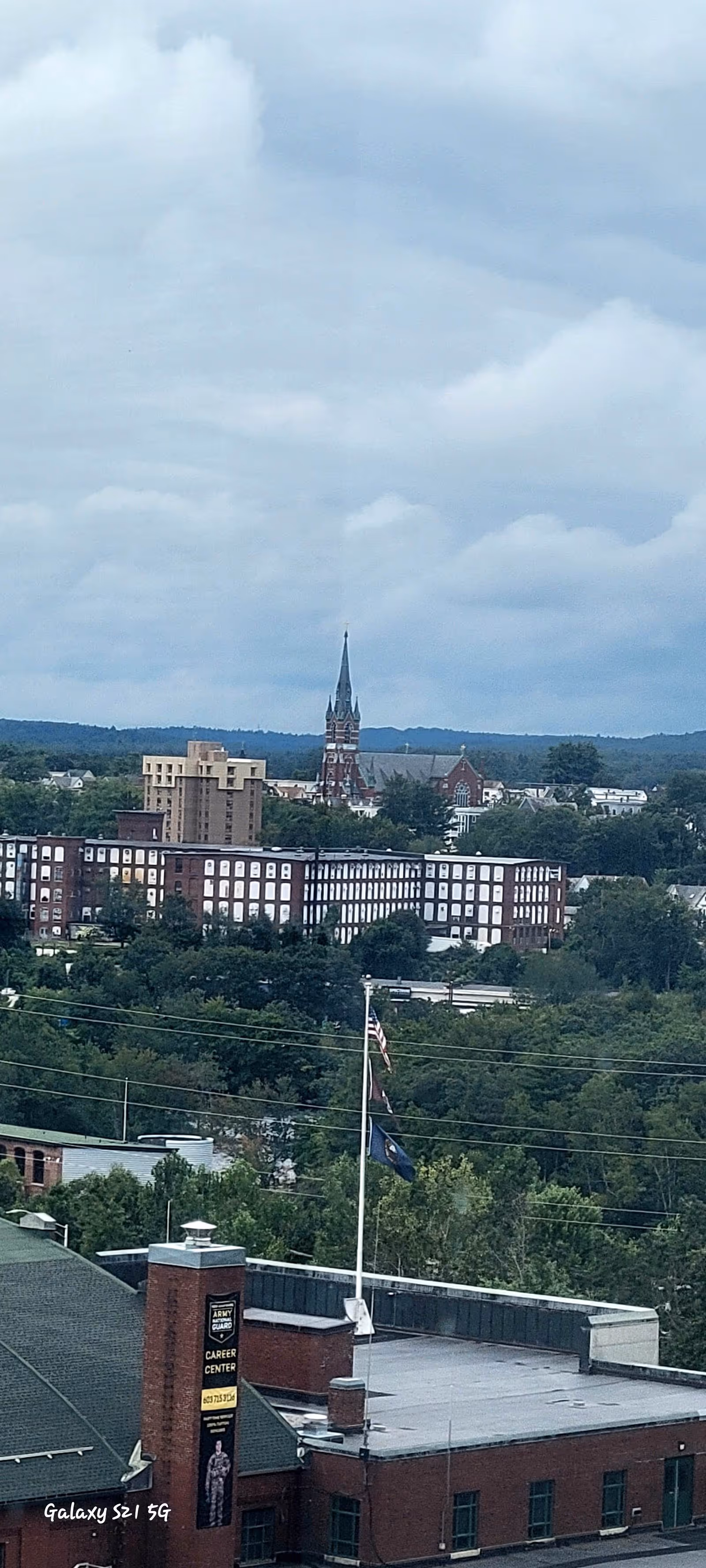 Urban cityscape featuring a church spire, trees, and a flag in the foreground.