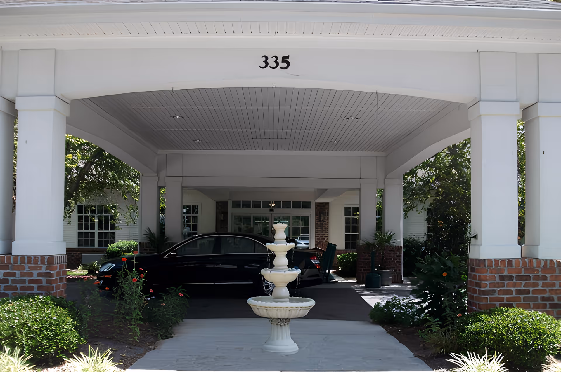 Covered entrance area of a building with the number 335 displayed above. A white three-tiered fountain is centered in the foreground, surrounded by green bushes and flowers. A black car is parked under the covered area near the entrance, with windows and doors visible in the background.