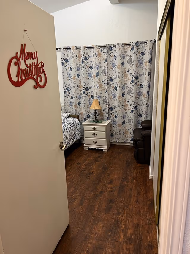 Bedroom with floral curtains, bed, nightstand, lamp, and recliner.