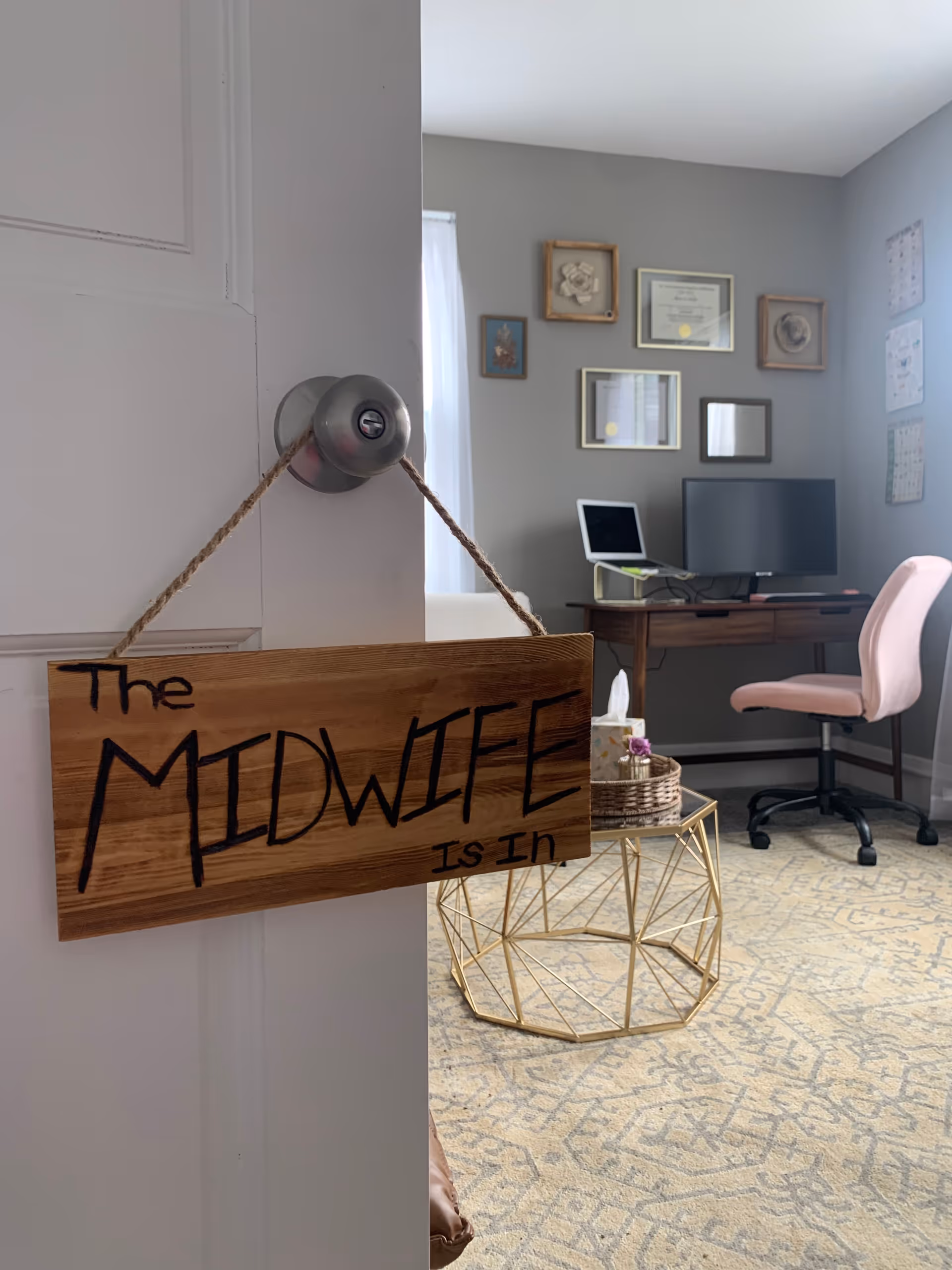 Door with wooden sign reading The MIDWIFE IS IN beside a desk in a lounge