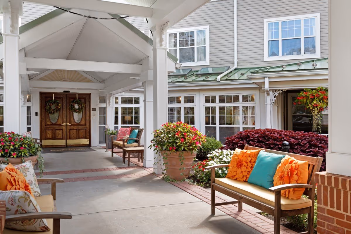 Covered entrance/porch of a senior living facility with benches, colorful cushions, potted plants, and double wooden doors.