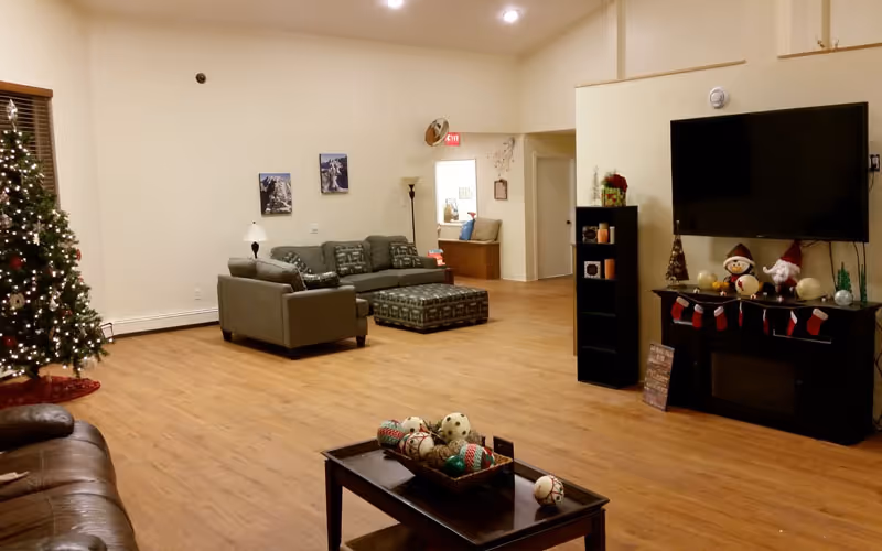 Spacious common room with sofas and ottoman, a wall-mounted TV, coffee table, and Christmas decorations including a tree and stockings.