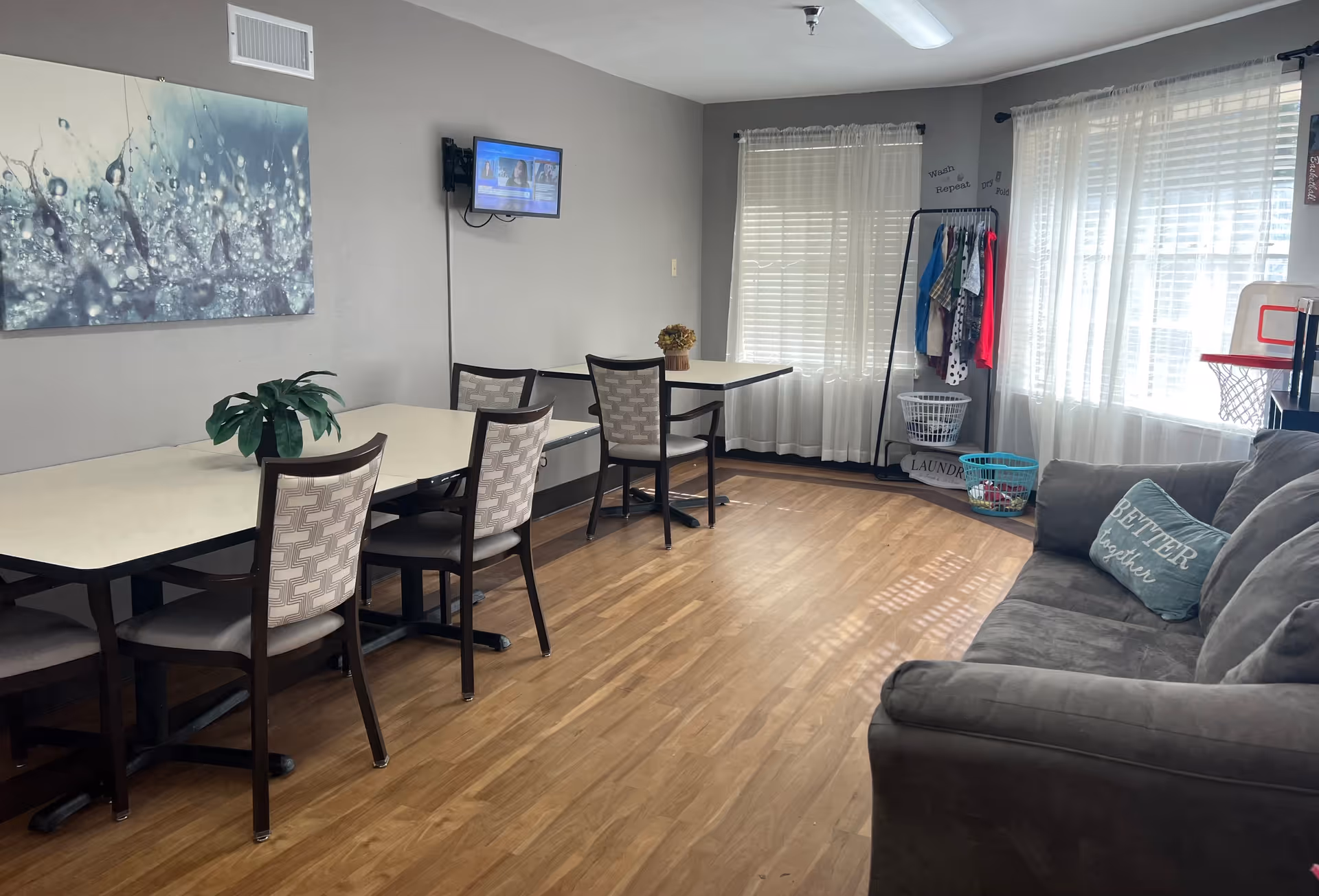 Bright communal lounge with dining tables and chairs, a gray sofa, a wall-mounted TV, and a small clothing rack by sunlit windows.