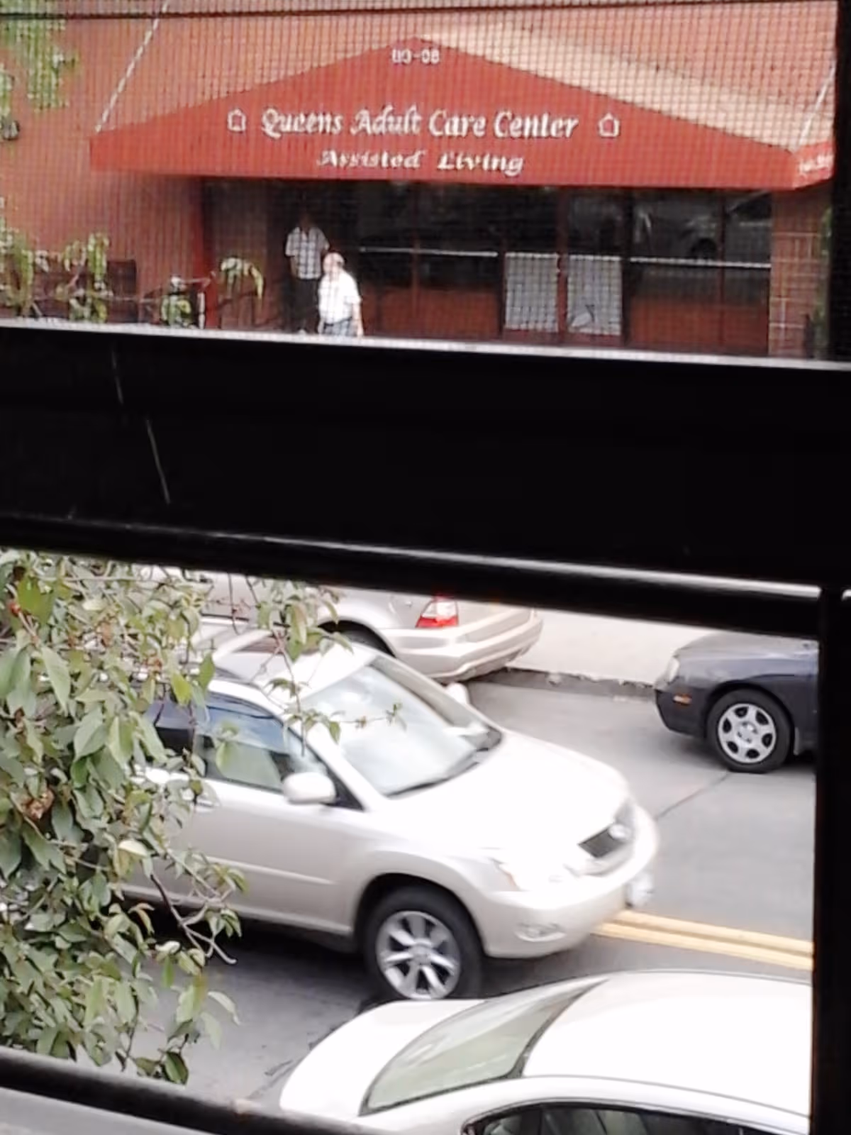 Exterior view of Queens Adult Care Center entrance with a red canopy and pedestrians near cars.