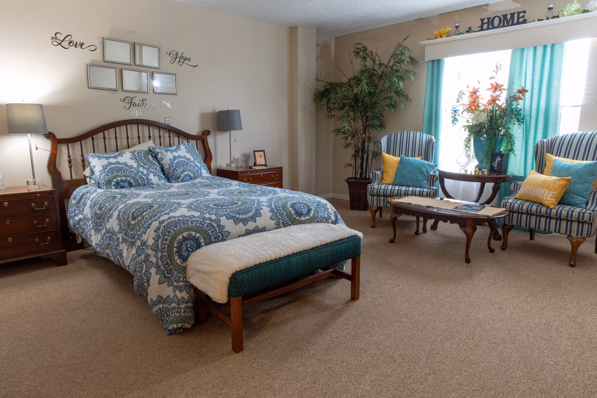 Well-decorated bedroom with a bed, nightstands, a bench at the foot, and a sitting area of two upholstered chairs by a window with plants.
