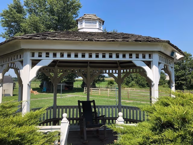 A white wooden gazebo with benches sits in a grassy outdoor area under blue skies.