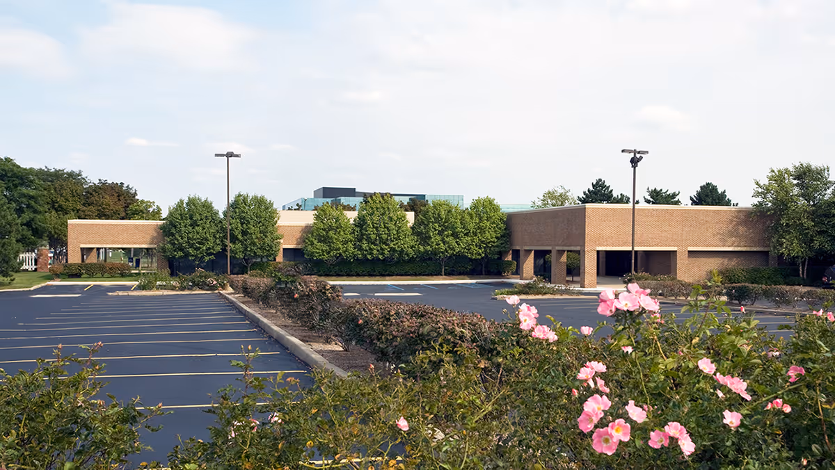 Brick senior living facility exterior with a large parking lot and blooming foreground flowers.