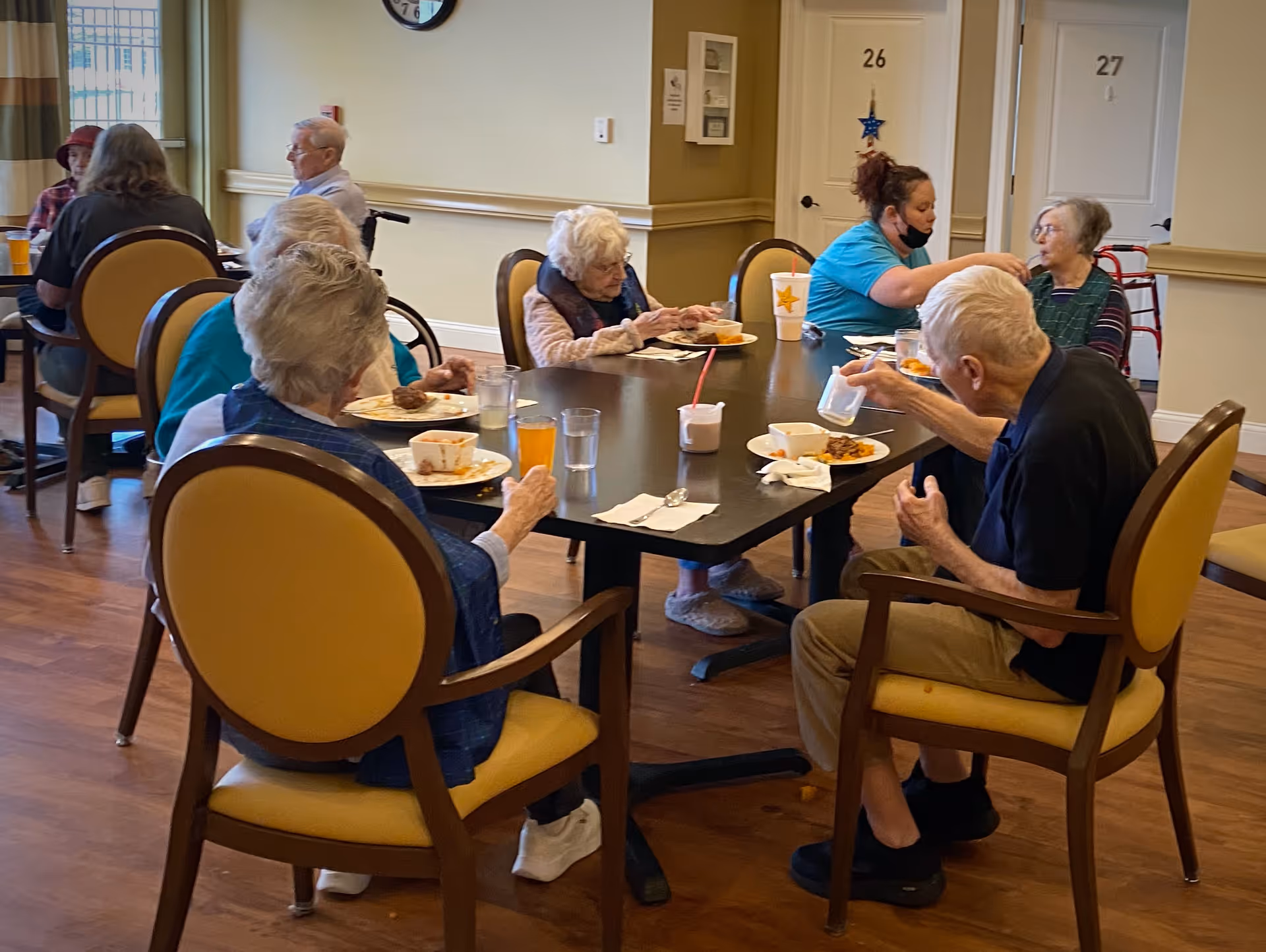 Several elderly individuals seated around a dining table in a communal dining area, eating and drinking. A caregiver wearing a blue shirt and a face mask is assisting one of the residents. The room has wooden floors, beige walls, and numbered doors in the background.