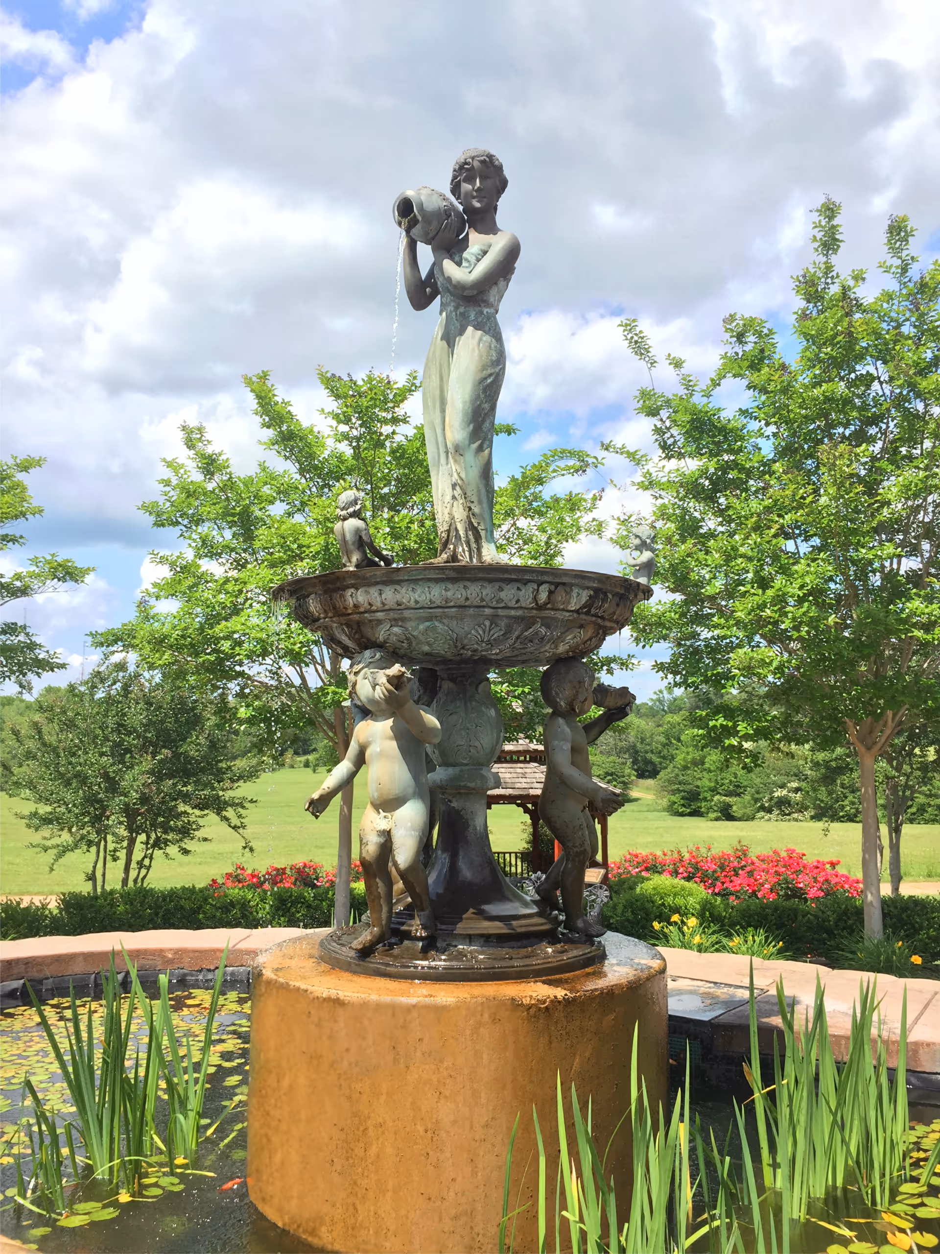 Bronze multi-figure fountain with a standing woman pouring water, amid trees and flowers