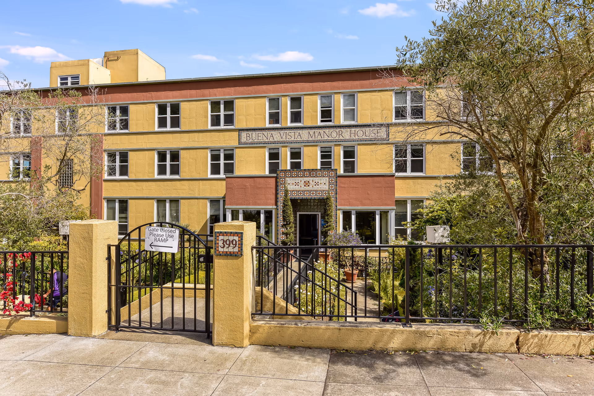 Exterior front view of Buena Vista Manor House, a three-story building with yellow and reddish-brown walls, multiple windows, and a gated entrance with a sign that reads 'Gate Closed Please Use Ramp'. The entrance is surrounded by greenery and plants.
