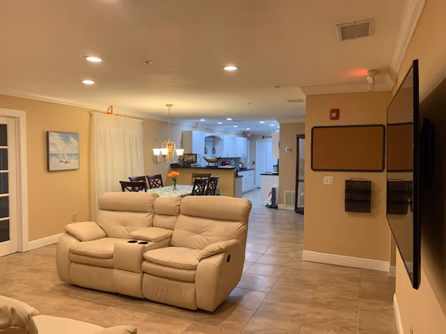 Interior view of a living area in an assisted living facility featuring a beige leather loveseat with cup holders, a dining table with chairs and a chandelier above it, and a kitchen area in the background with white cabinets and appliances. The walls are painted beige and the floor is tiled. A large flat-screen TV is mounted on the right wall.