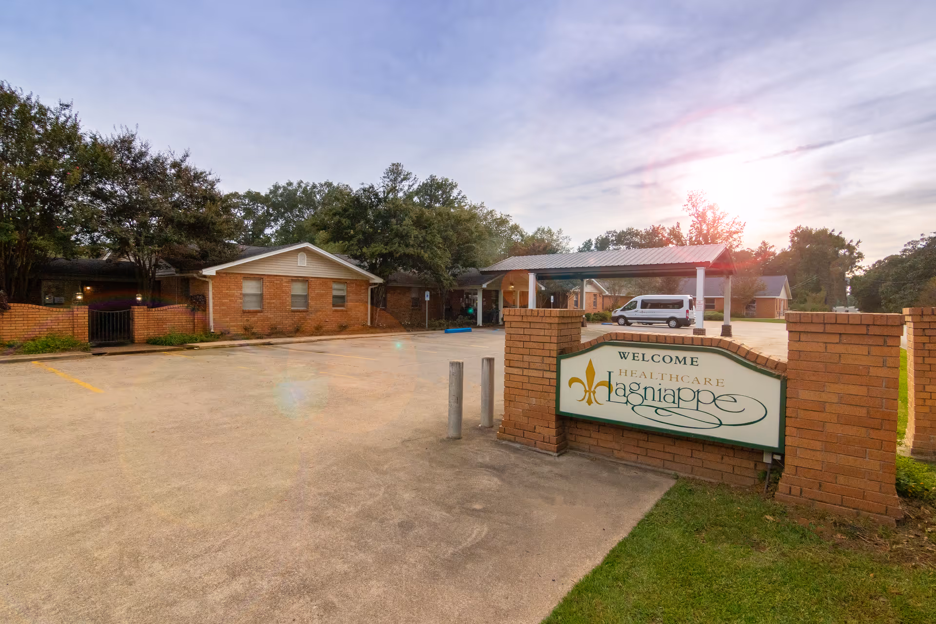 Exterior view of Lagniappe Health Care LLC facility showing a brick sign with the facility name, a parking lot, a covered entrance, and single-story brick buildings surrounded by trees under a partly cloudy sky with the sun low on the horizon.