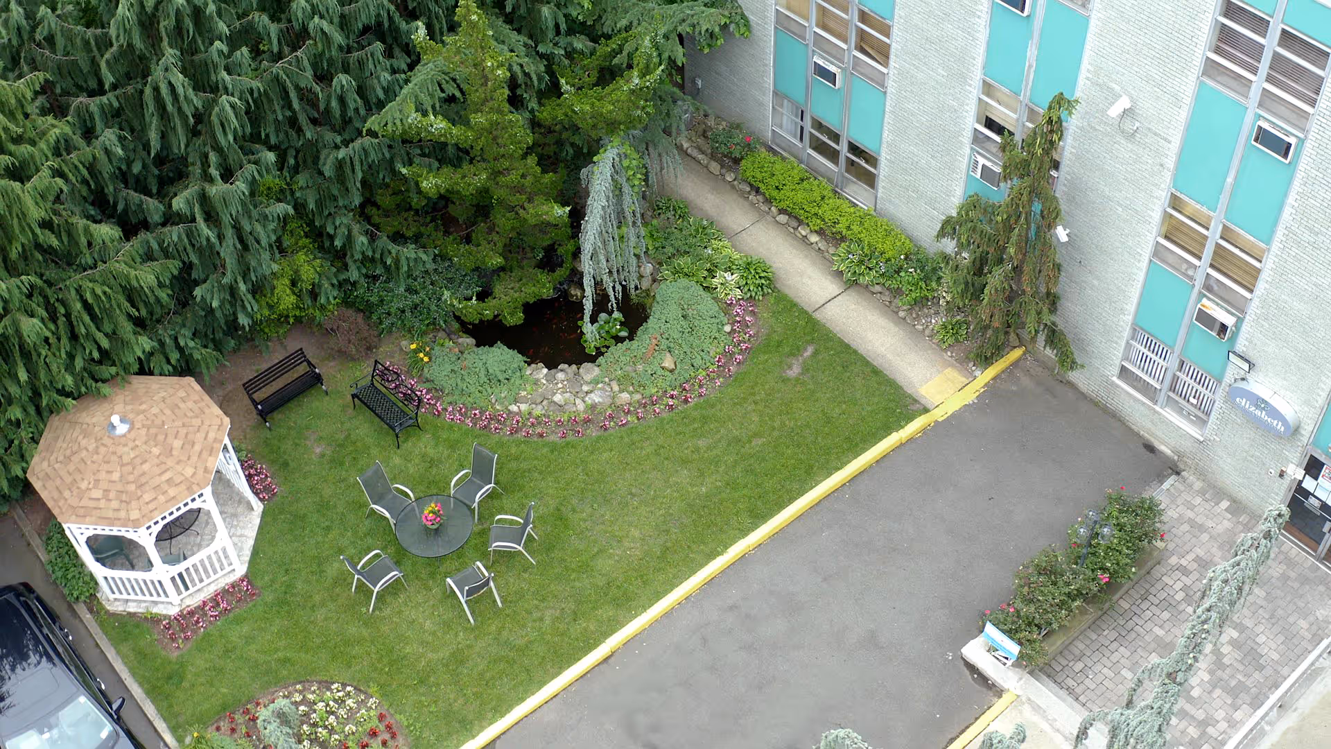 Aerial view of a garden area next to a building with a small gazebo, several chairs arranged around a circular table, benches, a small pond surrounded by flowers and greenery, and a paved pathway leading to the building entrance.