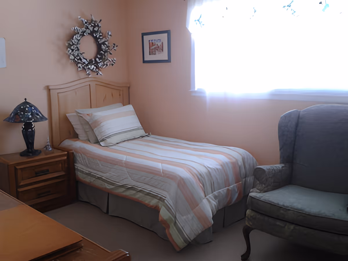 A small bedroom with a single bed covered in striped bedding, a wooden nightstand with a decorative lamp, a framed picture on the wall, a decorative wreath above the bed, and a cushioned armchair near a window with sheer curtains.