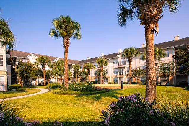 Sunlit courtyard of Brookdale Clear Lake with palm trees, trimmed lawn, and residential buildings.