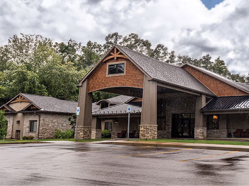 Senior living facility exterior with covered entry, stone pillars, and a parking lot.
