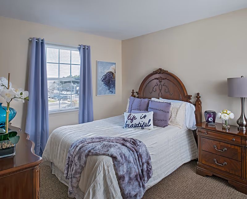 A furnished bedroom featuring a wooden headboard bed, lavender accents, and a matching nightstand.