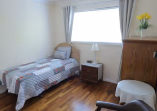 A small bedroom with a single bed covered with a patchwork quilt, a wooden nightstand with a lamp, a wooden dresser with a vase of yellow flowers, a round white table, and a chair. The room has hardwood flooring and a window with light gray curtains.