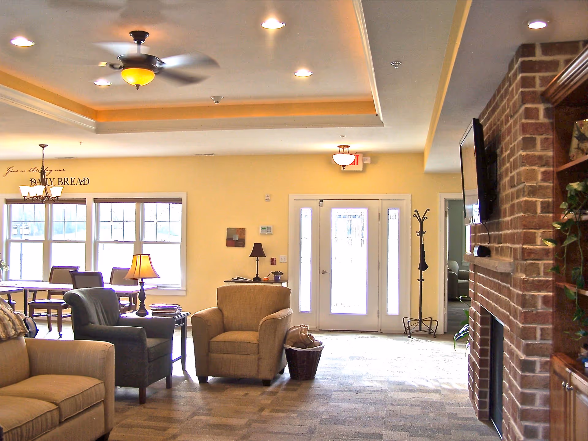 A cozy living room area in Rosewood Cottage featuring beige and dark armchairs, a beige sofa, a ceiling fan with light, a brick fireplace with a mounted TV, a coat rack, and a dining area with a table and chairs near large windows letting in natural light.