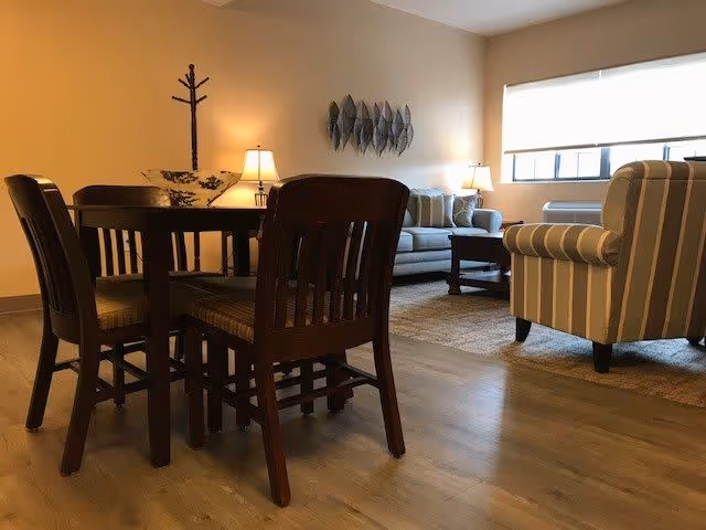 Cozy living area with a round wooden dining table and chairs in the foreground, a sofa, striped armchair, lamps, and wall decor near a window.