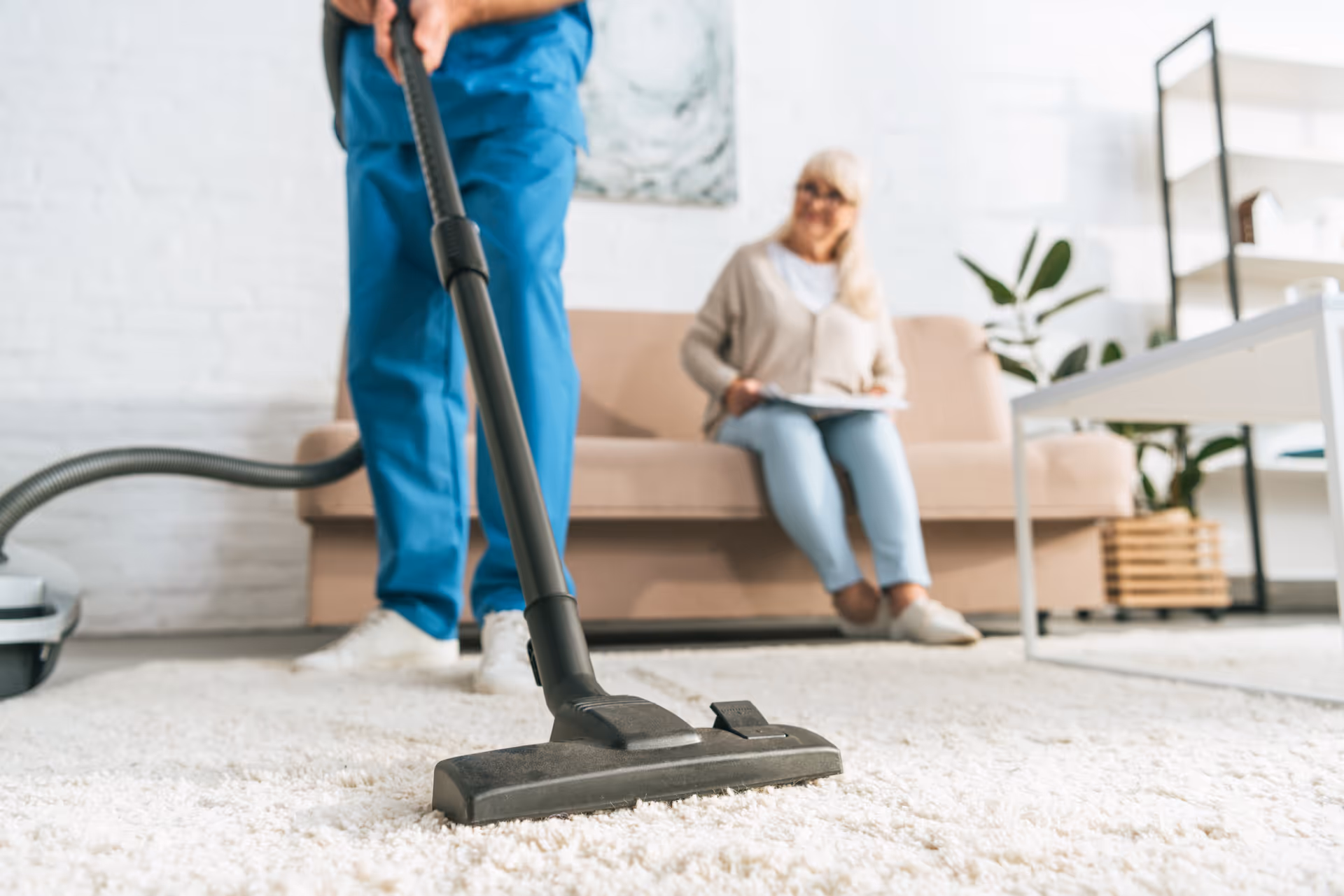 Caregiver vacuuming a living room while an elderly resident sits on the sofa.