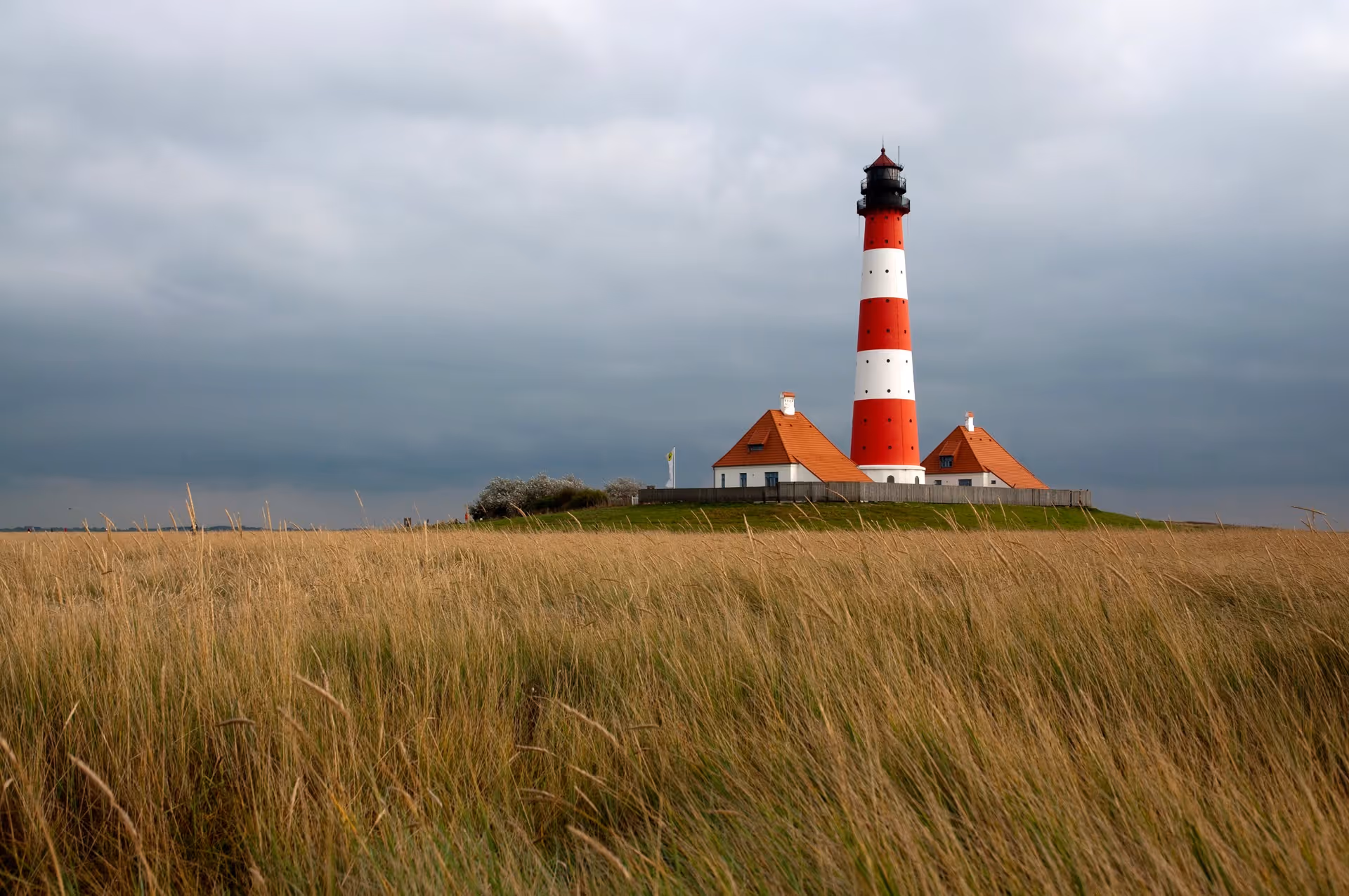 Red and white striped lighthouse with white buildings sits on a grassy field under cloudy skies.