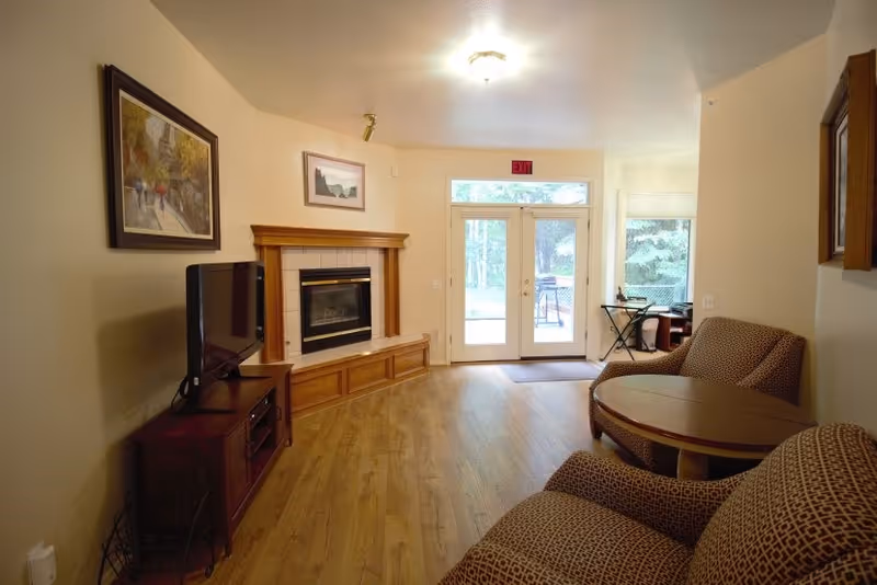 A cozy living room with wooden flooring, a fireplace with a wooden mantel, a flat-screen TV on a wooden stand, two patterned armchairs, a round wooden coffee table, framed artwork on the walls, and glass double doors leading outside to a patio area with trees visible through the doors.