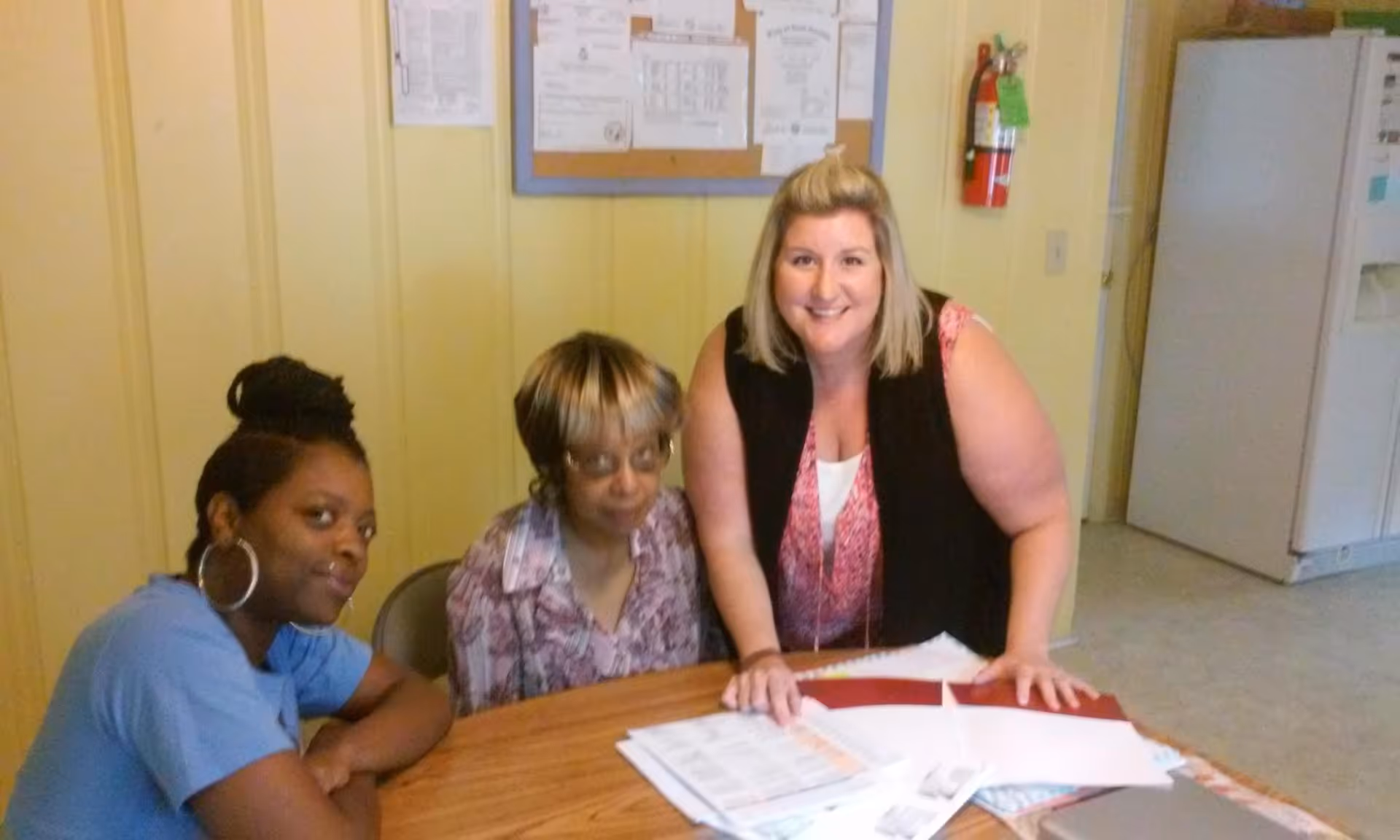 Three women sitting and standing around a wooden table with papers and folders in a room with yellow walls, a bulletin board, a fire extinguisher, and a refrigerator in the background.