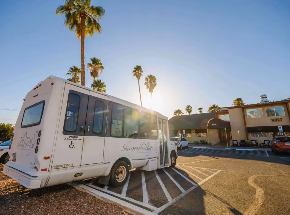 A white shuttle bus with Sherwood Village Assisted Living & Memory Care branding is parked in a designated handicap parking space outside a two-story assisted living facility building with palm trees in the background and the sun low in the sky.