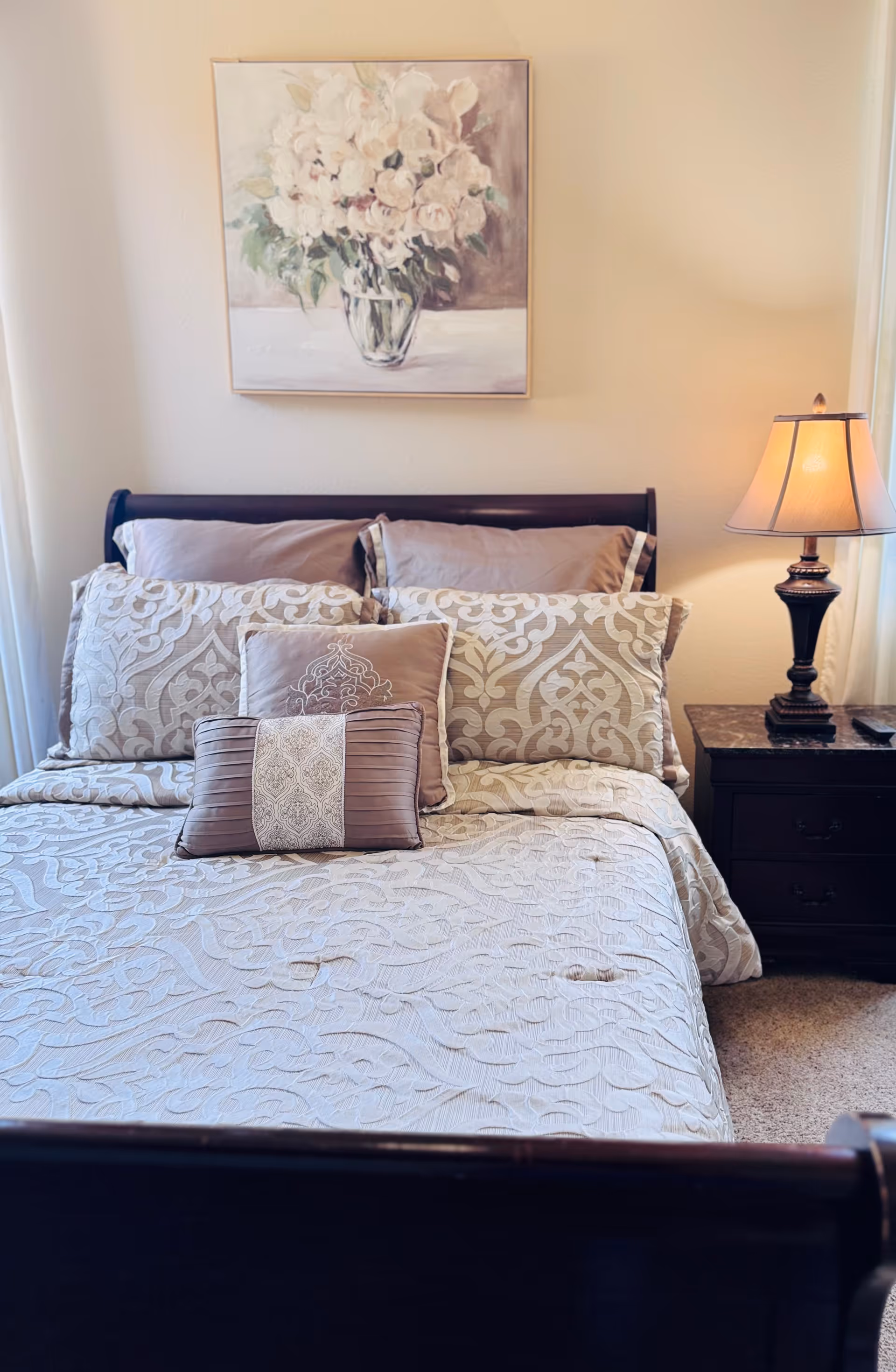 A neatly made bedroom with a bed, decorative pillows, nightstand, lamp, and floral wall art.