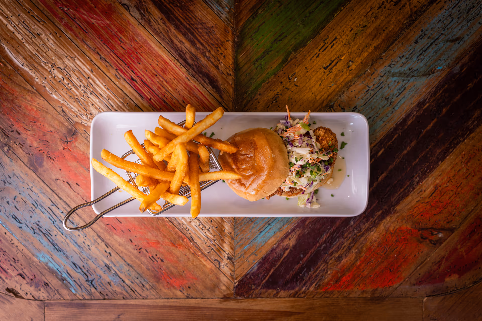 A rectangular white plate with a serving of French fries in a small metal basket and a sandwich with coleslaw on a rustic wooden table with colorful, weathered planks.