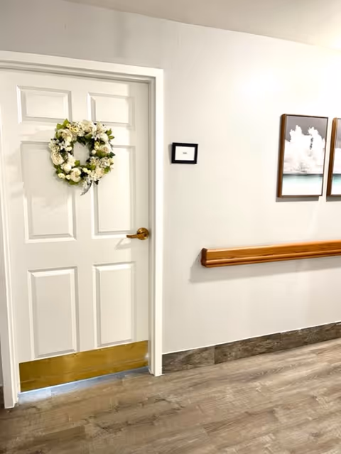 Interior hallway of a senior living facility with a white door decorated with a floral wreath, a wooden handrail on the wall, two framed pictures, and wood-look flooring.