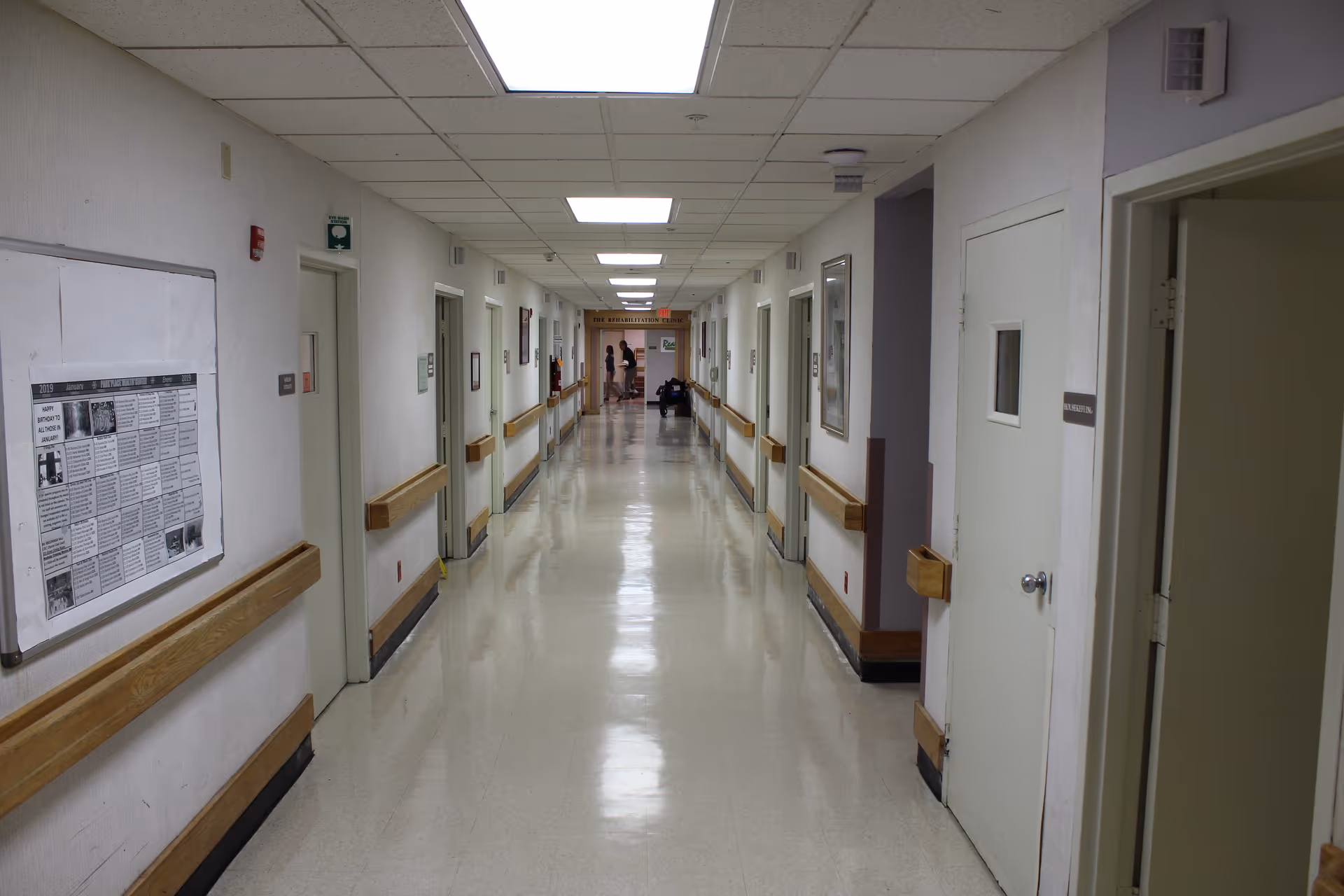 A long, clean hallway in a care facility with white walls, beige floor, and wooden handrails on both sides. Several closed doors line the hallway, and a bulletin board is visible on the left wall. Two people are seen standing and talking at the far end of the corridor.