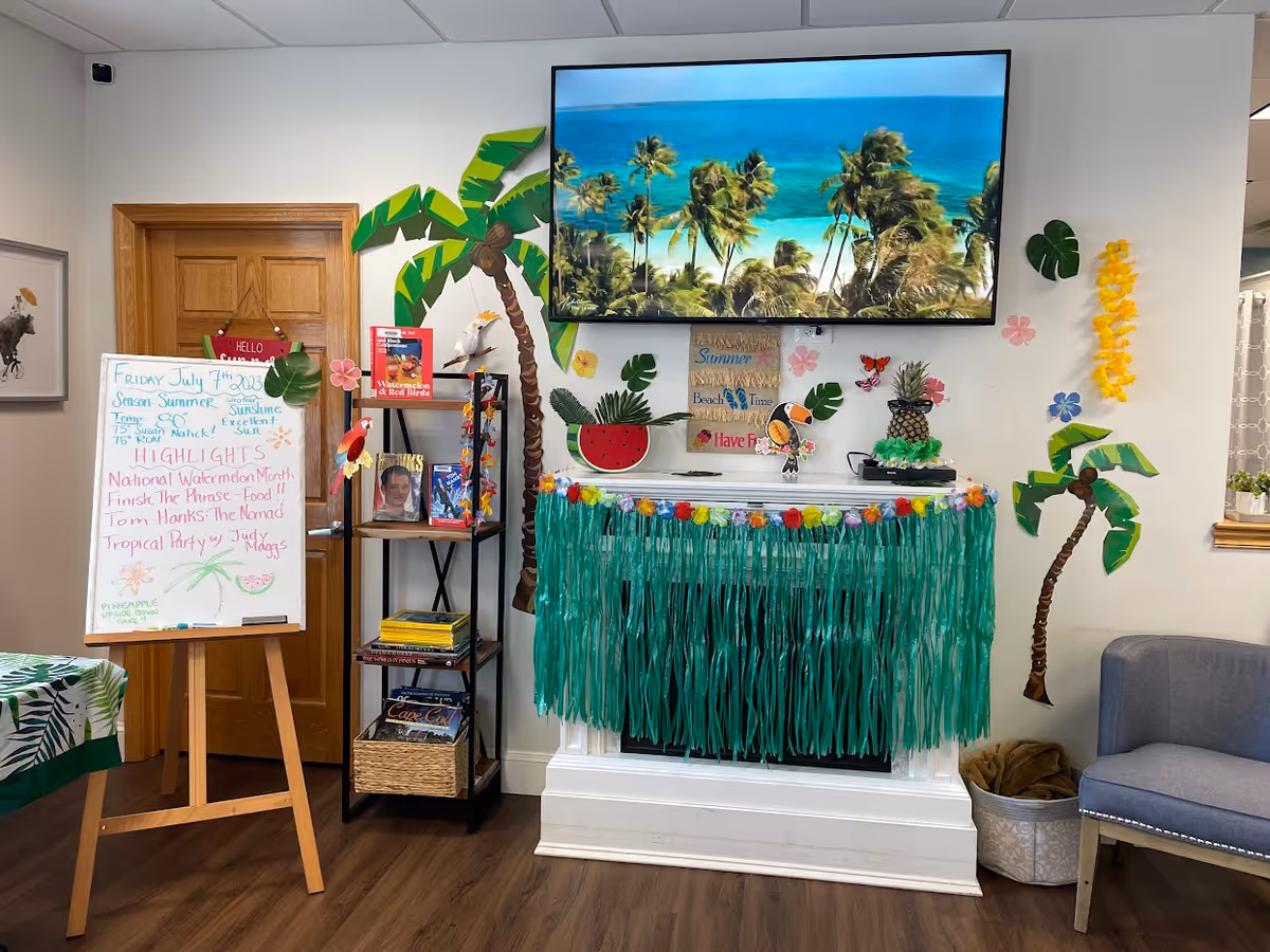 Bright activity room at Elder-Well Adult Day Program with tropical decor, whiteboard, shelves, and TV.