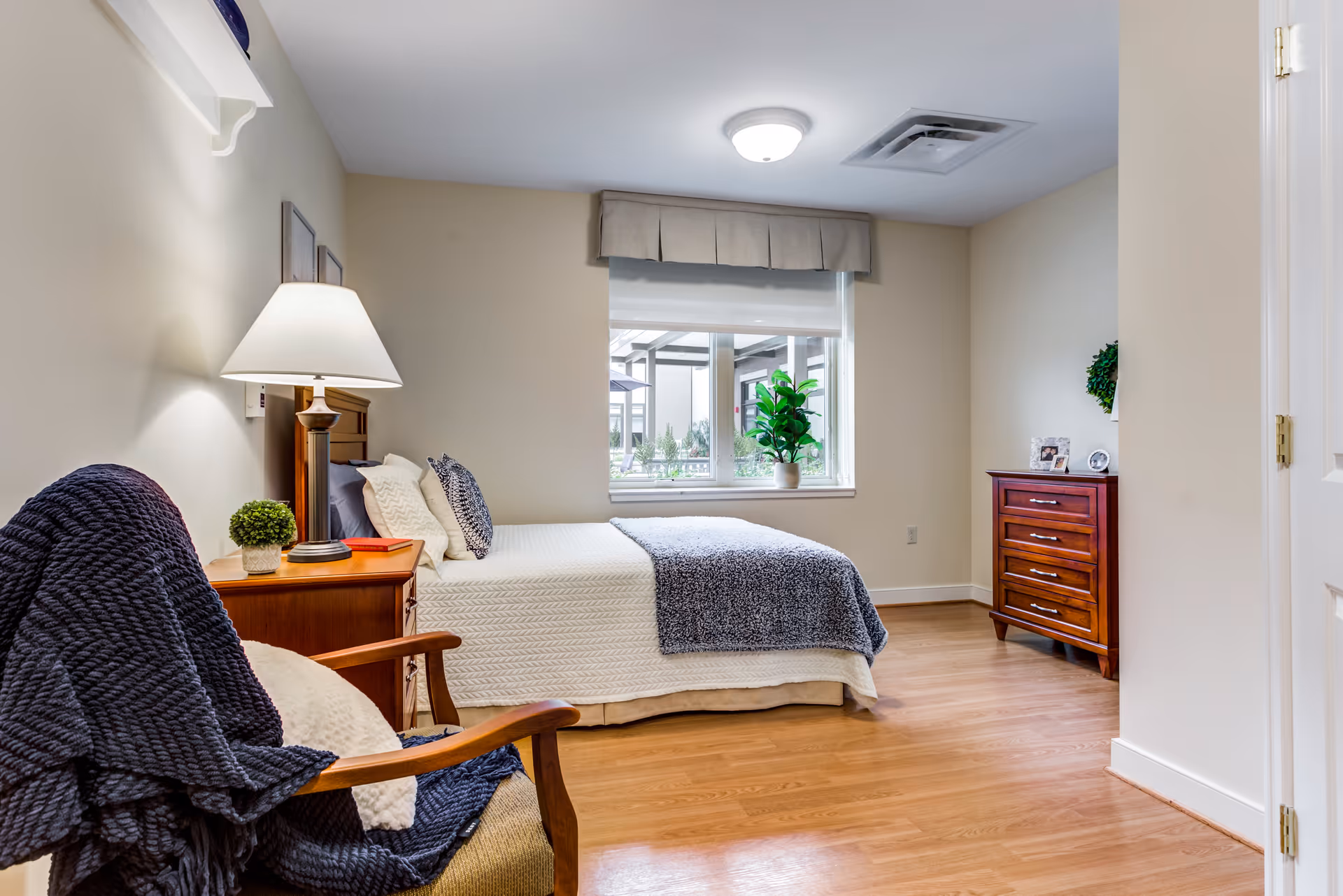 A cozy bedroom in a senior living facility featuring a neatly made bed with white and gray bedding, a wooden nightstand with a lamp and a small plant, a wooden armchair draped with a dark gray blanket, a wooden dresser with framed photos and a small plant, and a window with a beige valance letting in natural light.