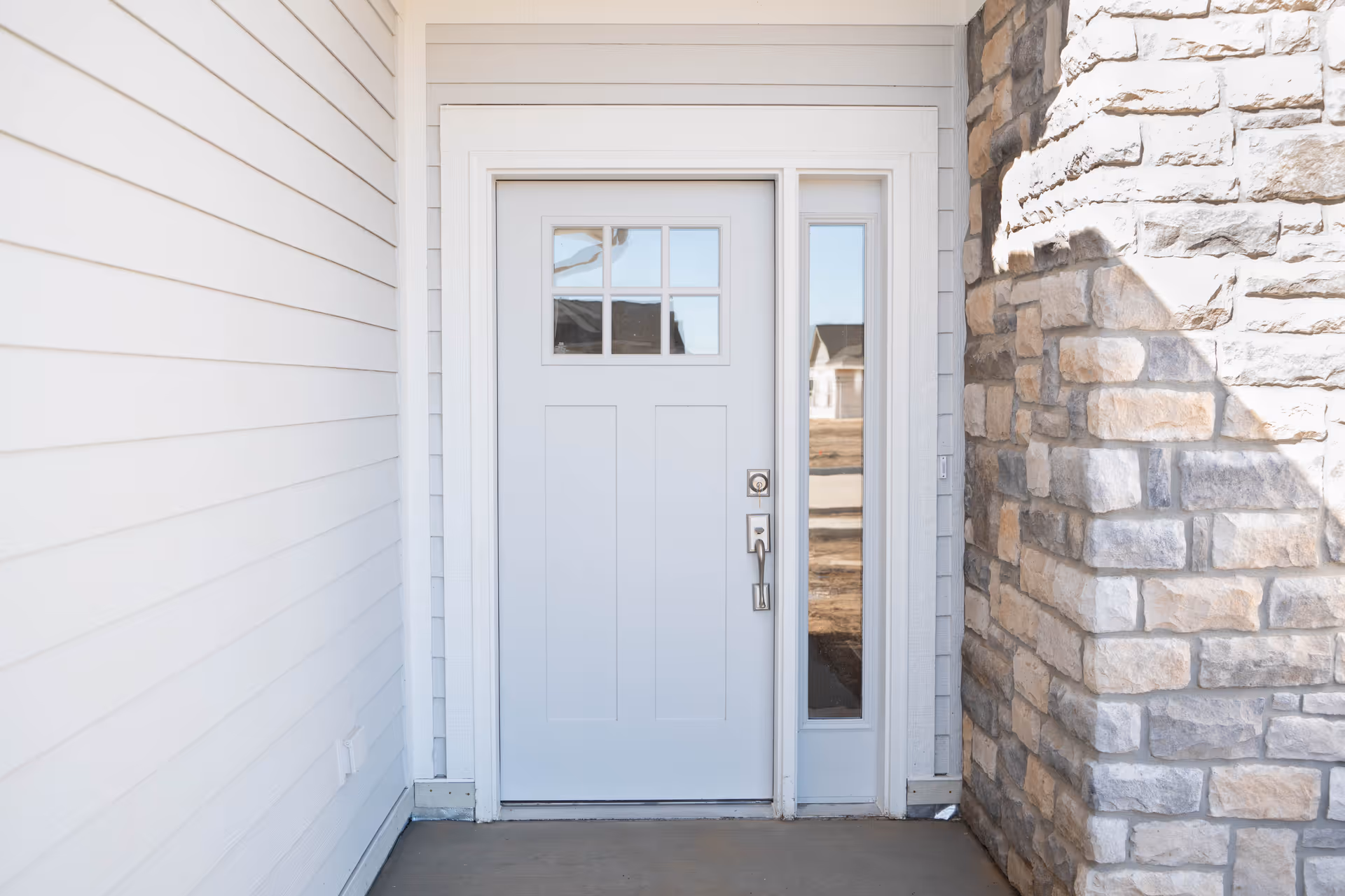 A white front door with a small window panel at the top and a narrow vertical window on the right side. The door is framed by white siding on the left and a stone wall on the right.