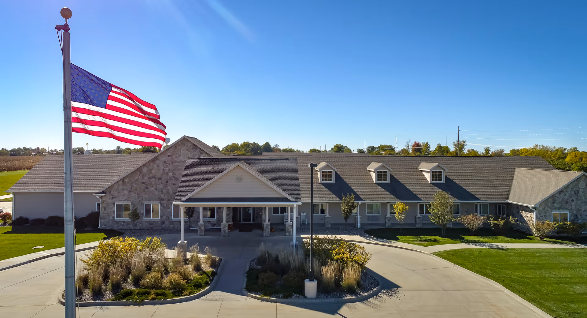Front exterior view of a single-story assisted living and memory care facility with a stone and siding facade, a covered entrance, landscaped greenery, and an American flag flying on a flagpole in front.