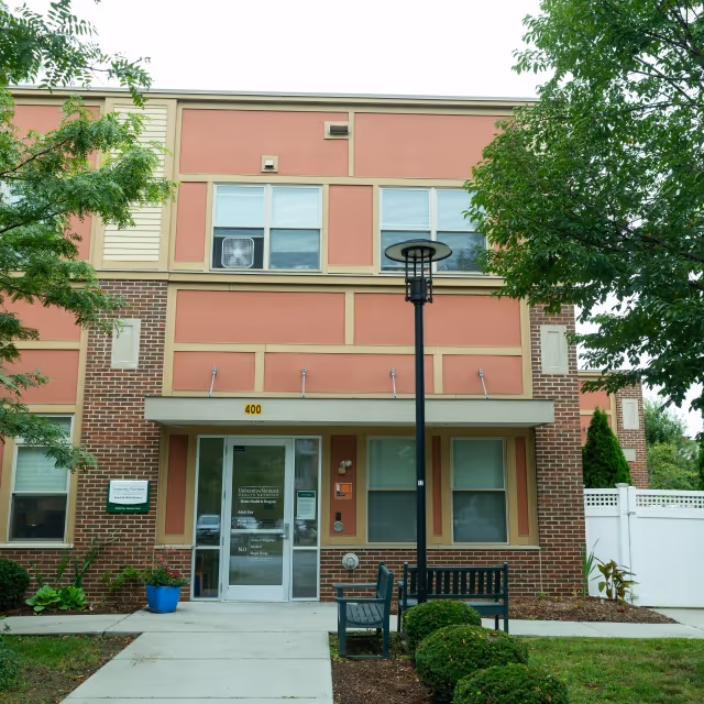 Front of Adult Day Program facility with pink panels, glass doors, bench, and greenery.
