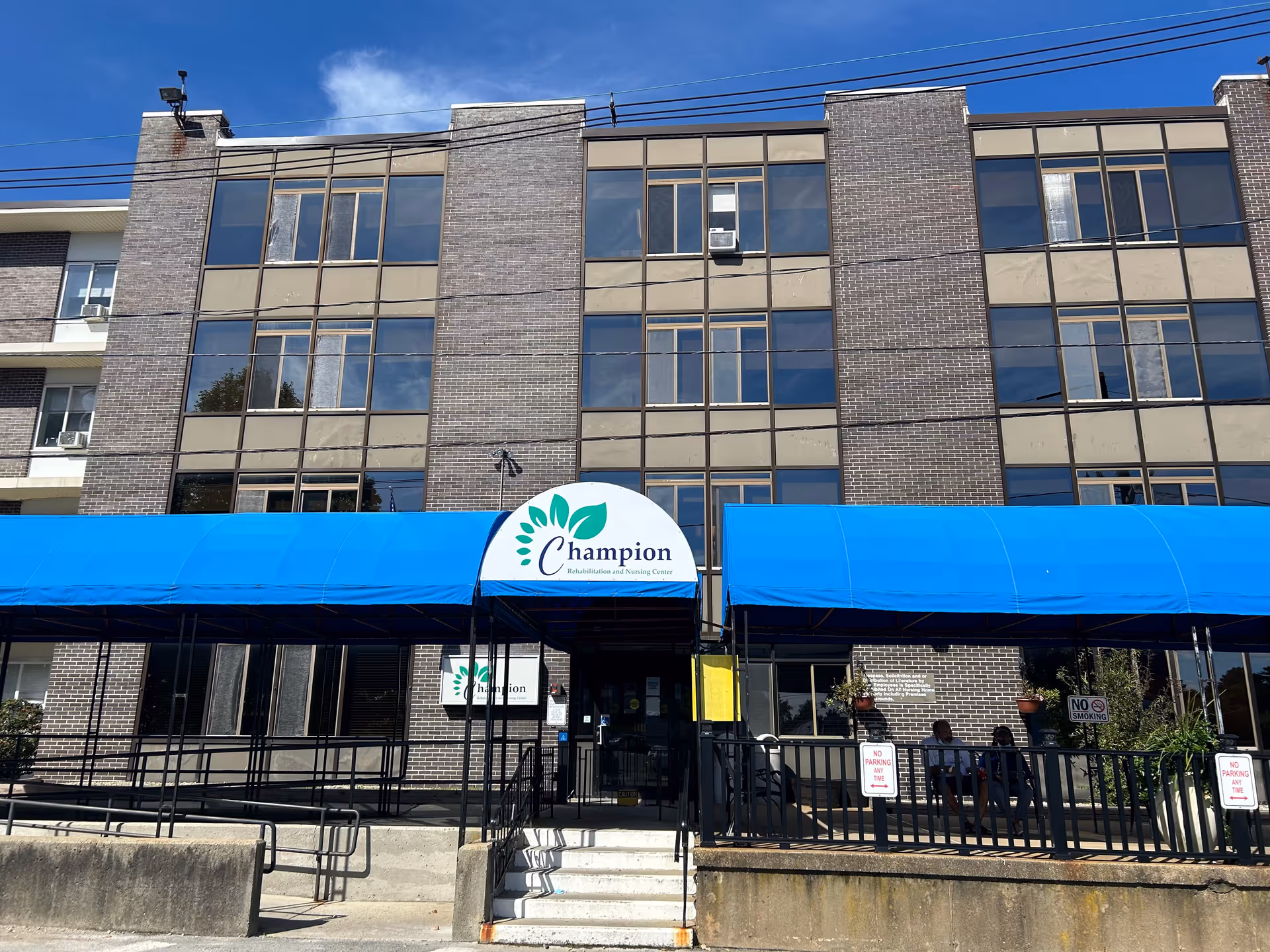 Front exterior view of a multi-story brick building with large windows and blue awnings over the entrance. A sign above the entrance reads 'Champion Rehabilitation and Nursing Center'. There are railings and a small set of stairs leading to the entrance, with a few people sitting on the right side under the awning.