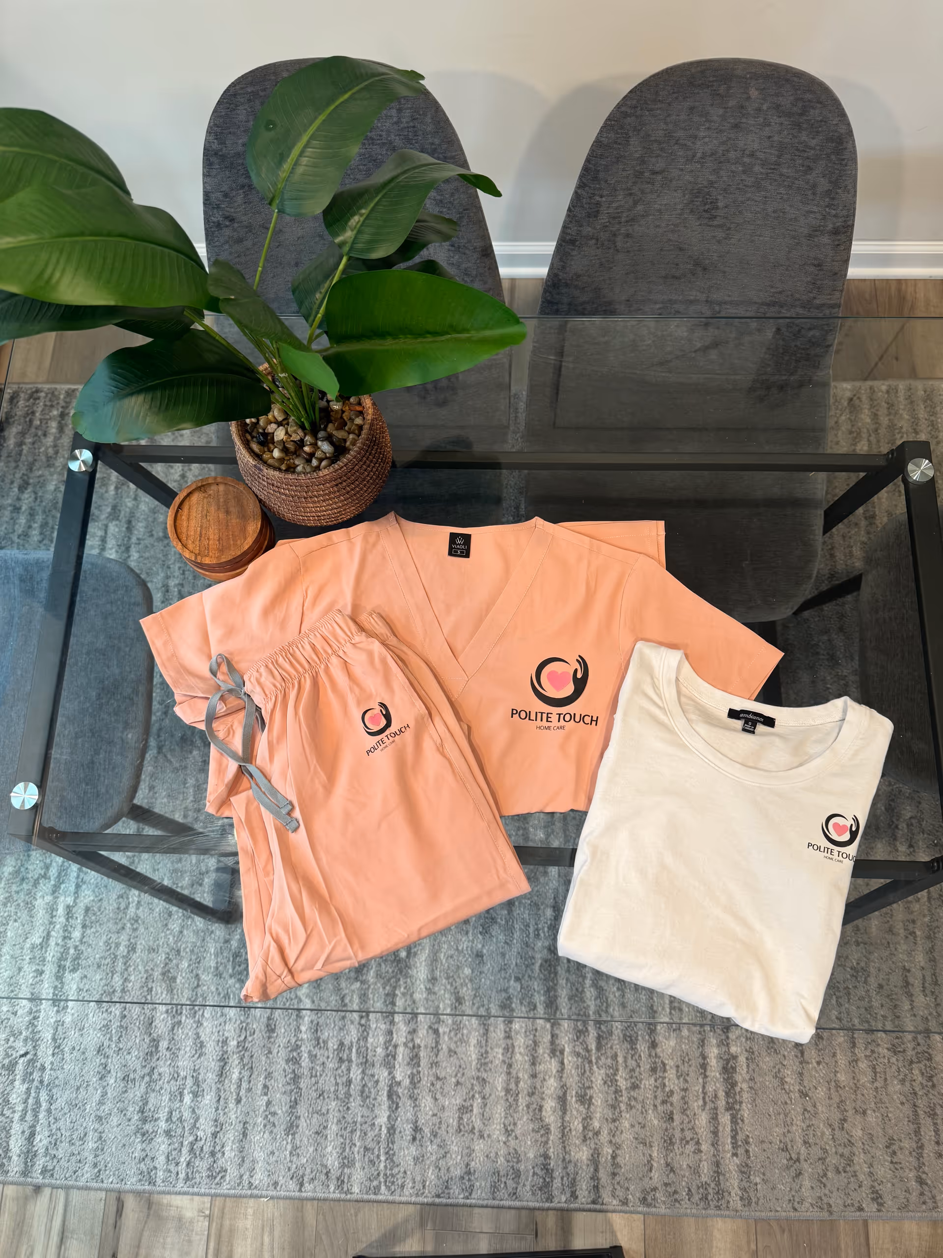 Pink scrubs and a beige shirt with Polite Touch logo on a glass coffee table