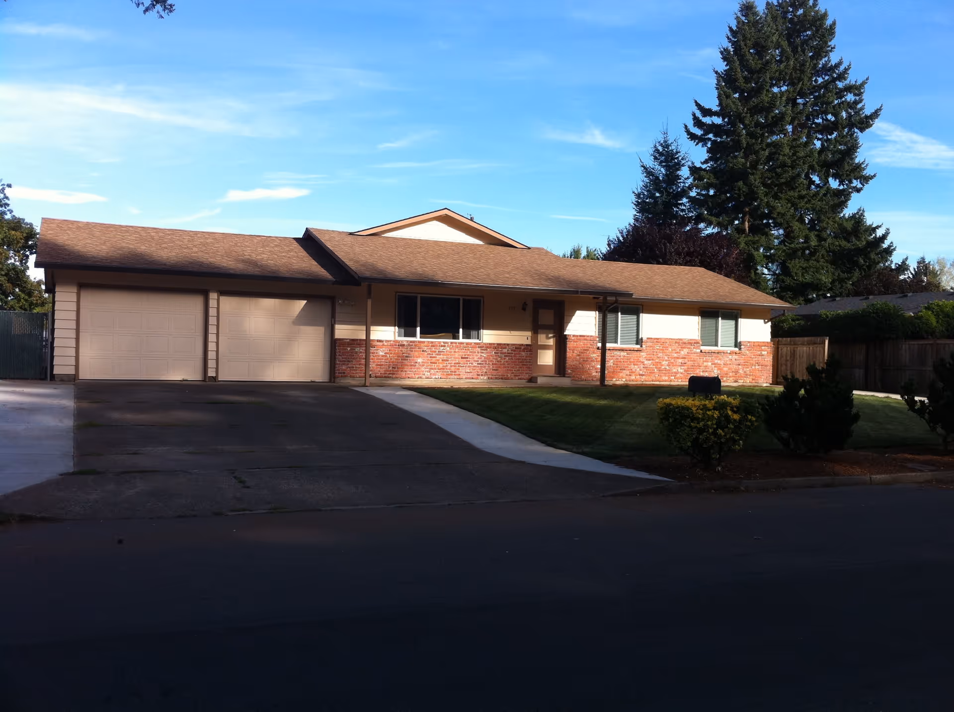 Single-story residential building with a brown roof, beige siding, and red brick accents. The house has two garage doors on the left side, a central window, and a front door with a small porch. The front yard is landscaped with green grass, bushes, and a large tree in the background under a clear blue sky.