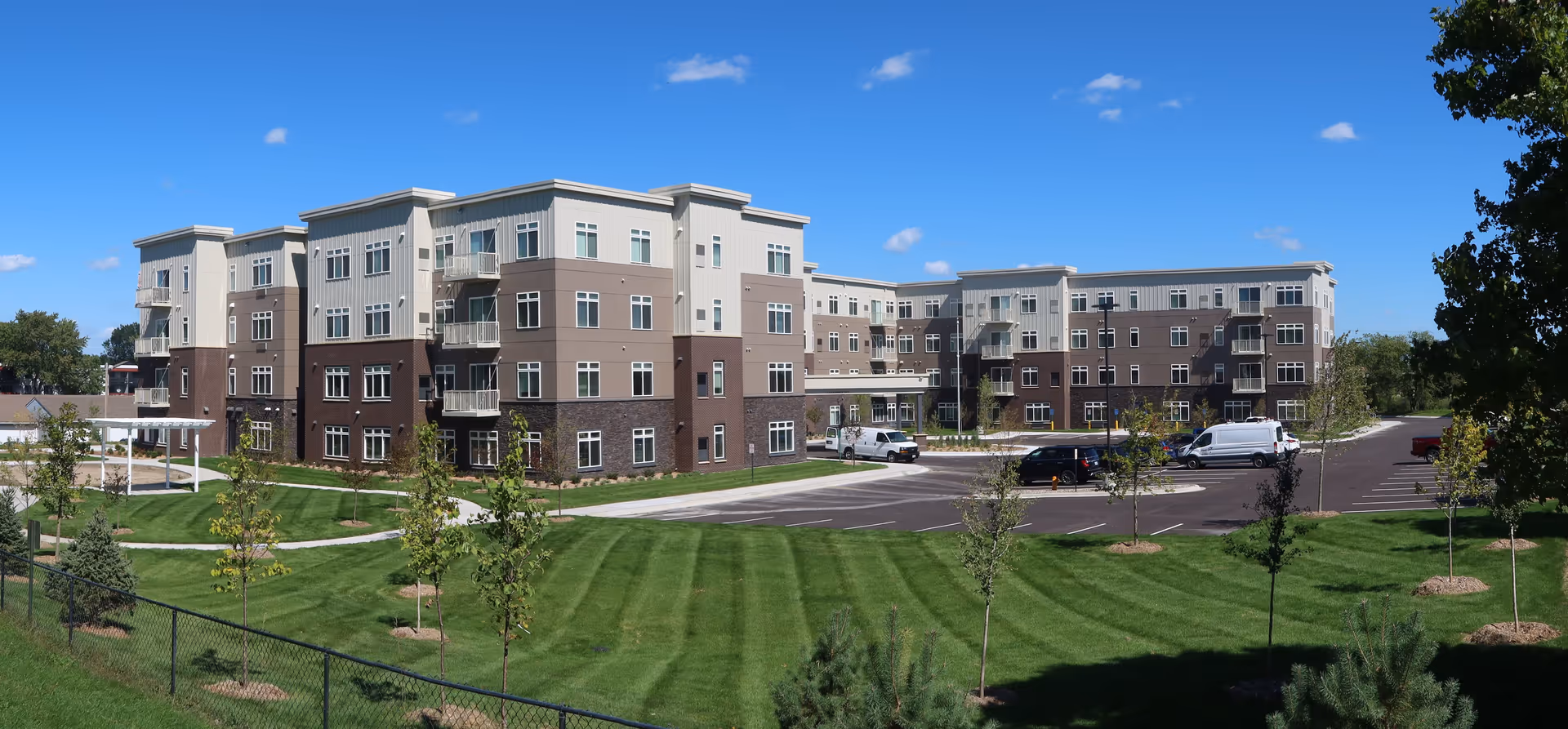 Panoramic exterior view of a four-story senior living building with a parking lot, manicured lawn, and a clear blue sky.