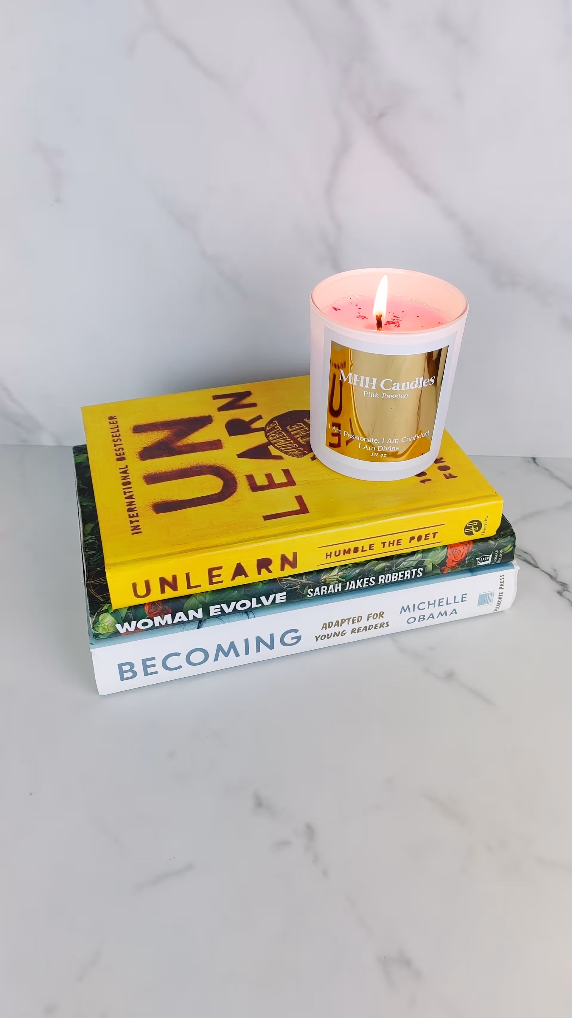 Lit pink candle labeled MHH Candles atop a stack of books on marble surface.