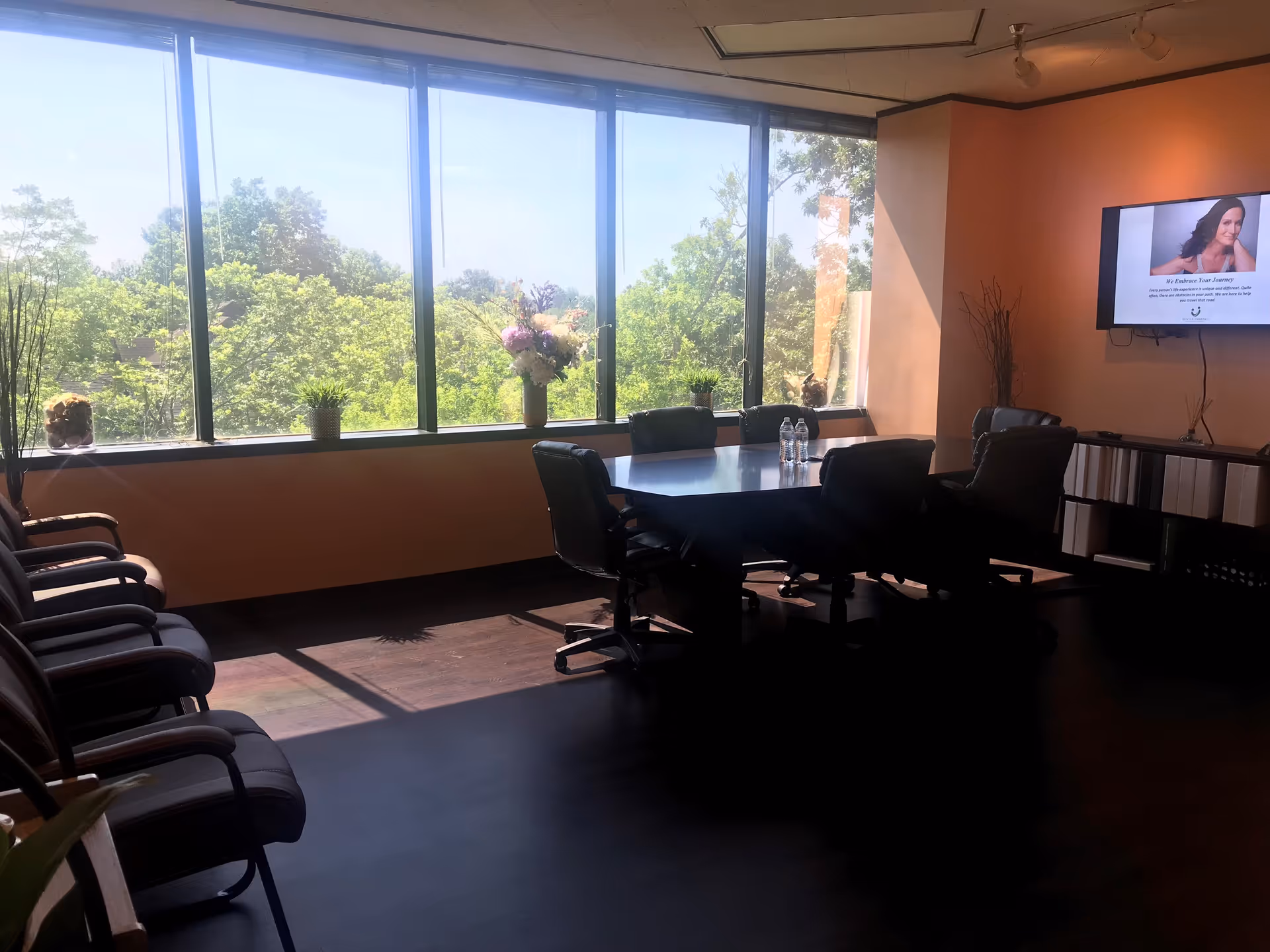 Sunlit conference room with a long table, chairs, large windows, and a TV.
